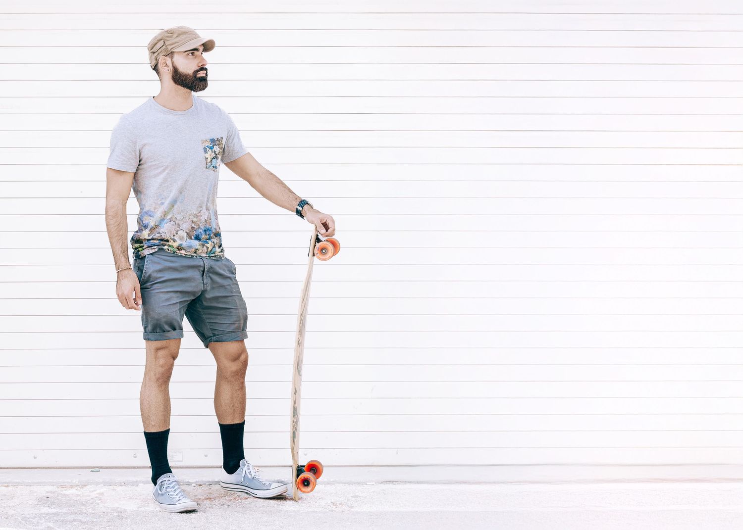 Casual hipster with beard stands against white wall holding longboard while wearing gray t-shirt and denim shorts.