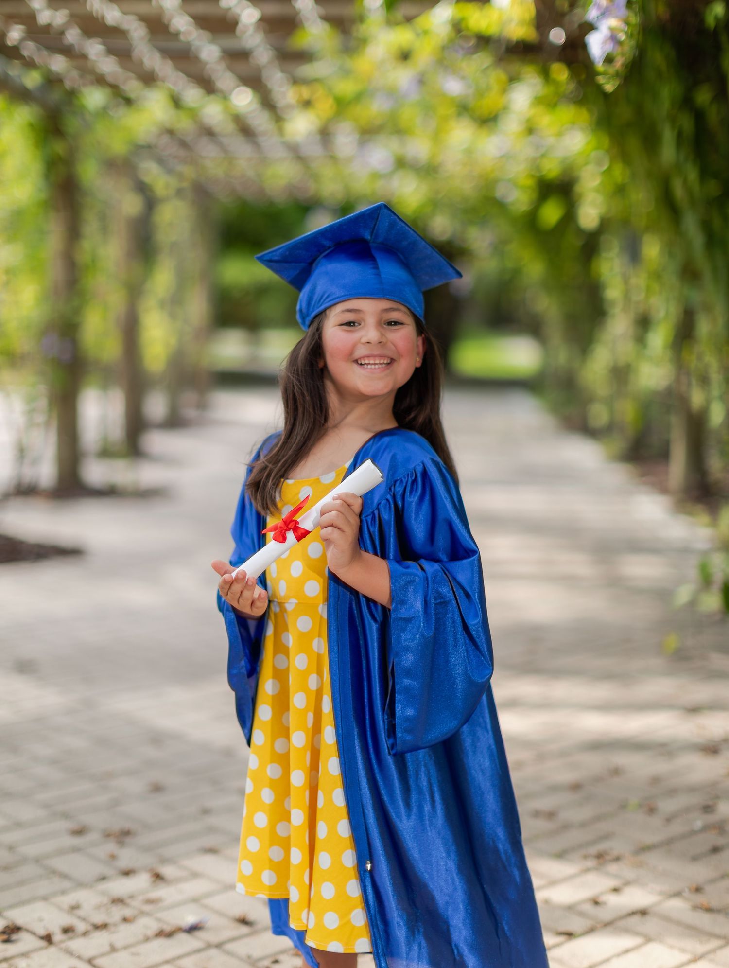Graduate in bright blue cap and gown stands smiling on garden path wearing yellow polka dot dress underneath.