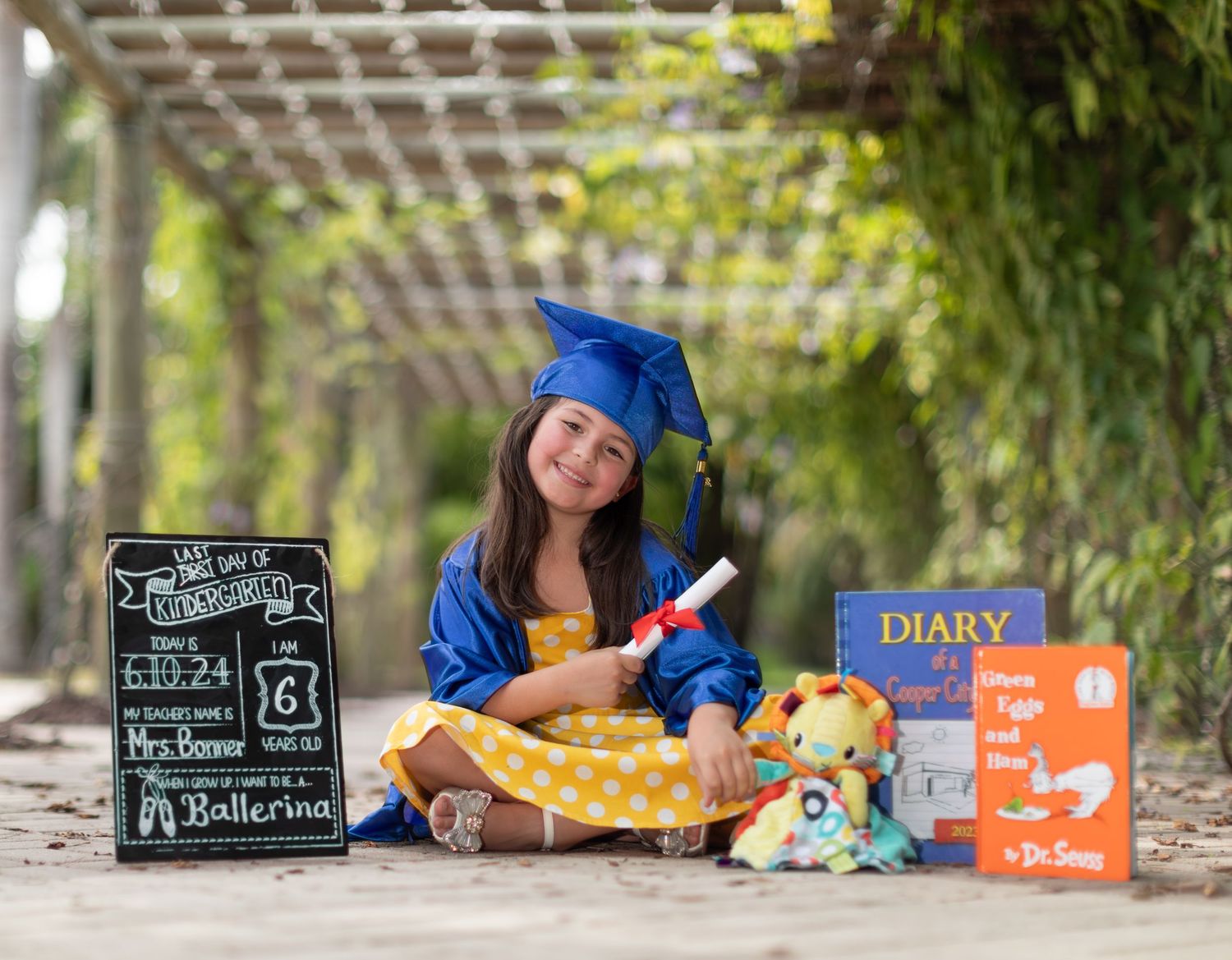 A graduate in blue cap and gown sits outdoors with books and stuffed animals for a graduation photo session.