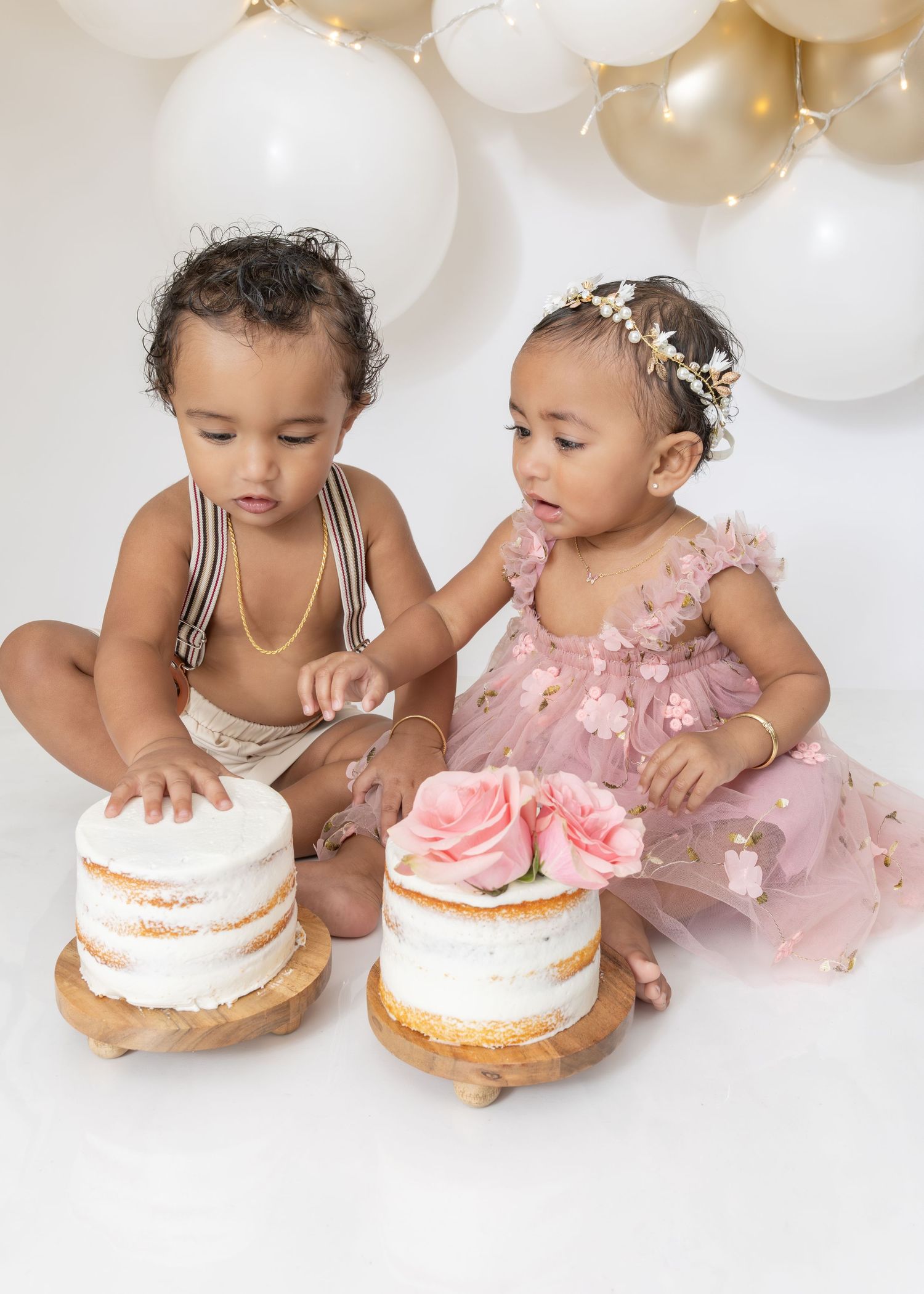 Two babies in white and pink outfits sit together with naked cakes decorated with roses and white balloon decorations.