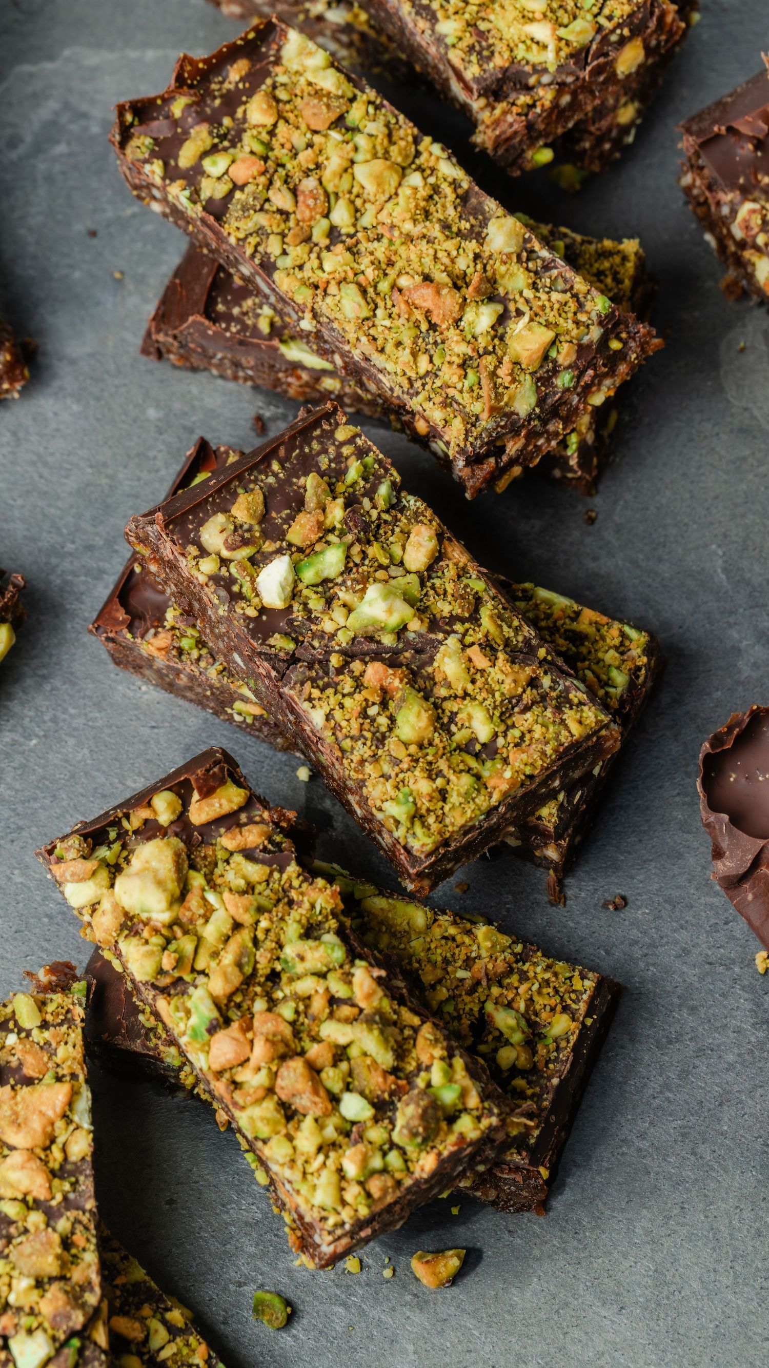 Chocolate pistachio bars topped with crushed nuts arranged diagonally on dark slate surface.