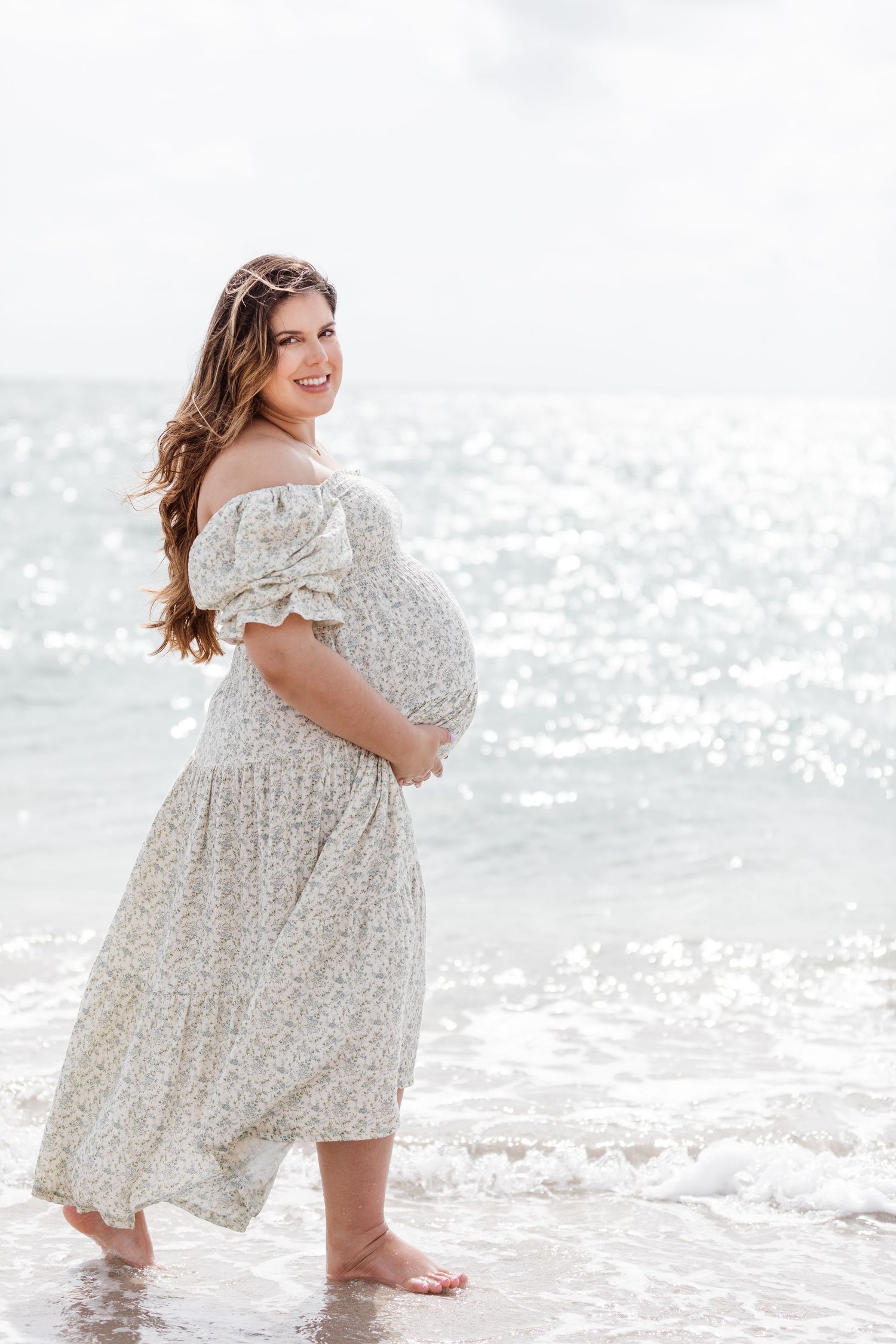 An expectant person in a flowy white floral dress stands barefoot at the ocean's edge on a bright sunny day.