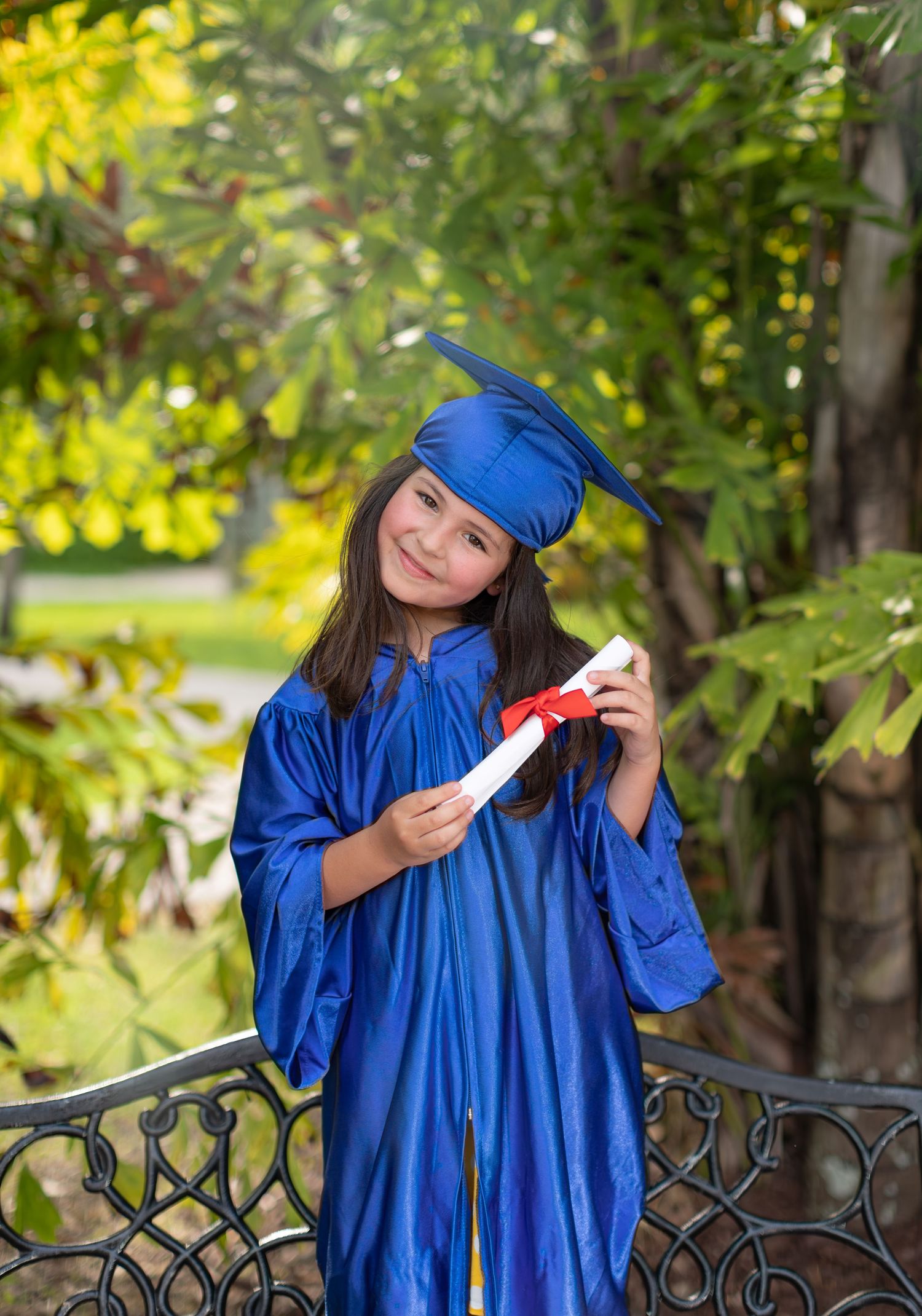 Graduate in bright blue cap and gown holds diploma while posing on garden bench with lush greenery in background.