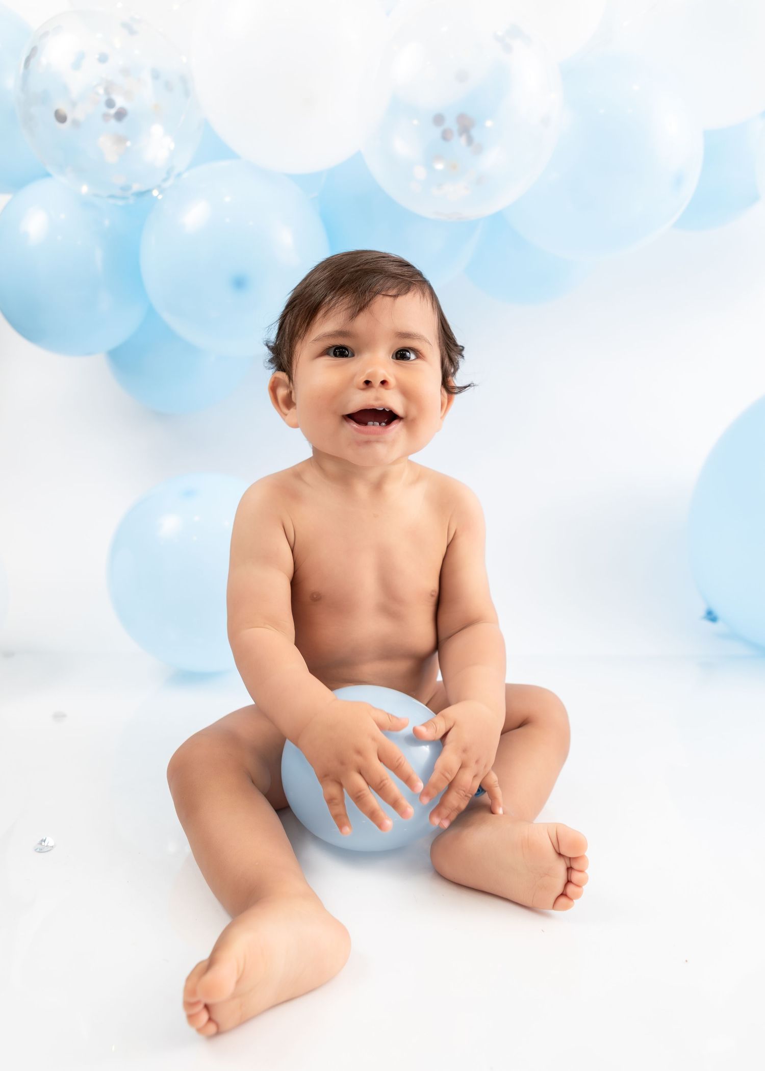 Happy baby sitting with blue balloon against white and blue balloon backdrop.