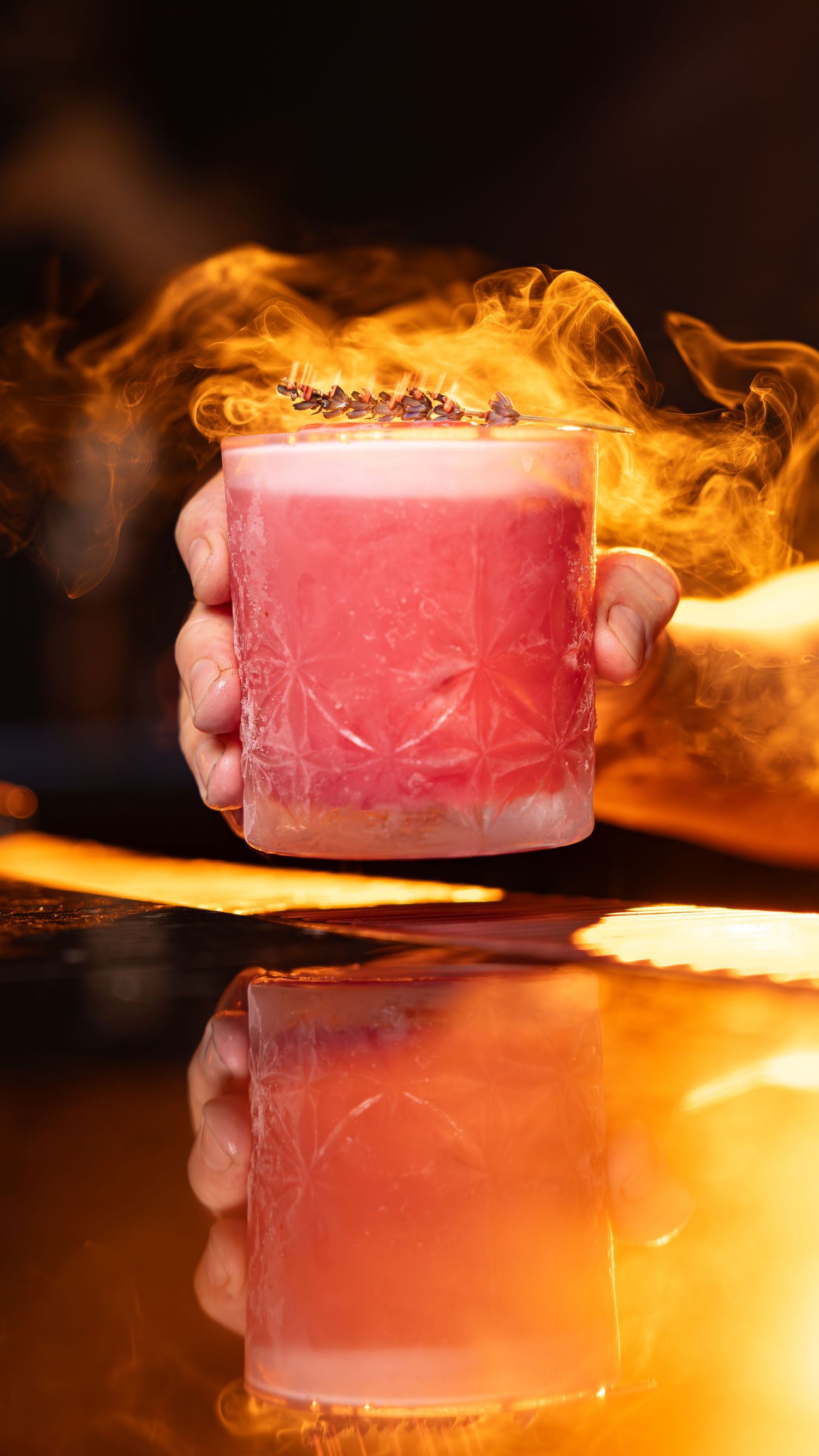 A dramatic cocktail with smoke and flames rising from a pink drink served in a crystal glass on a reflective surface.