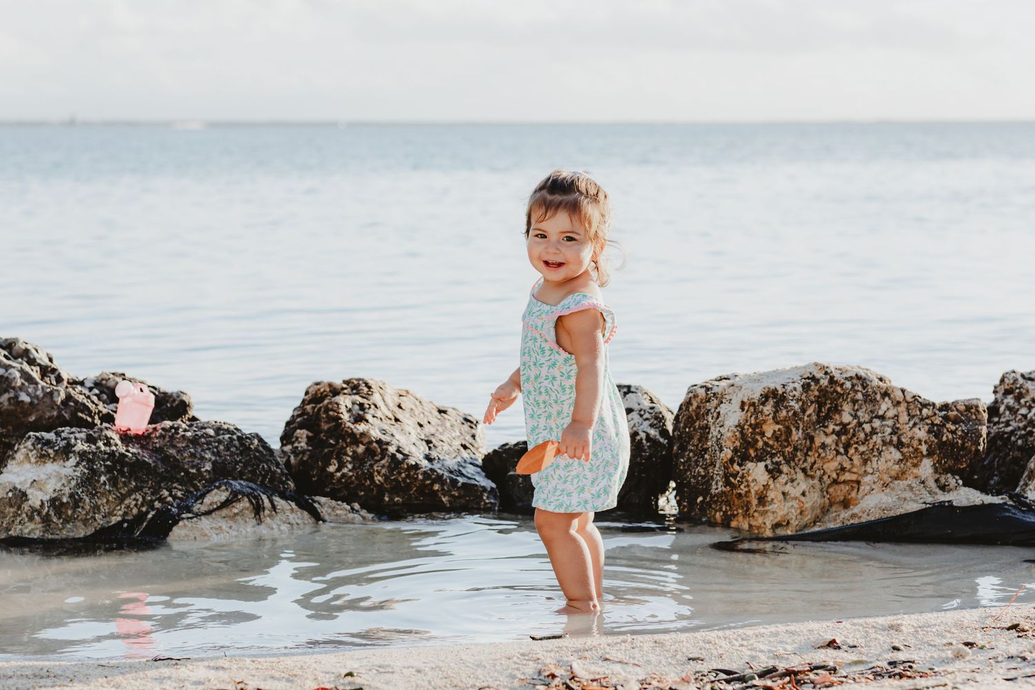 A small child plays at the edge of the water near large rocks on a beach at sunset.