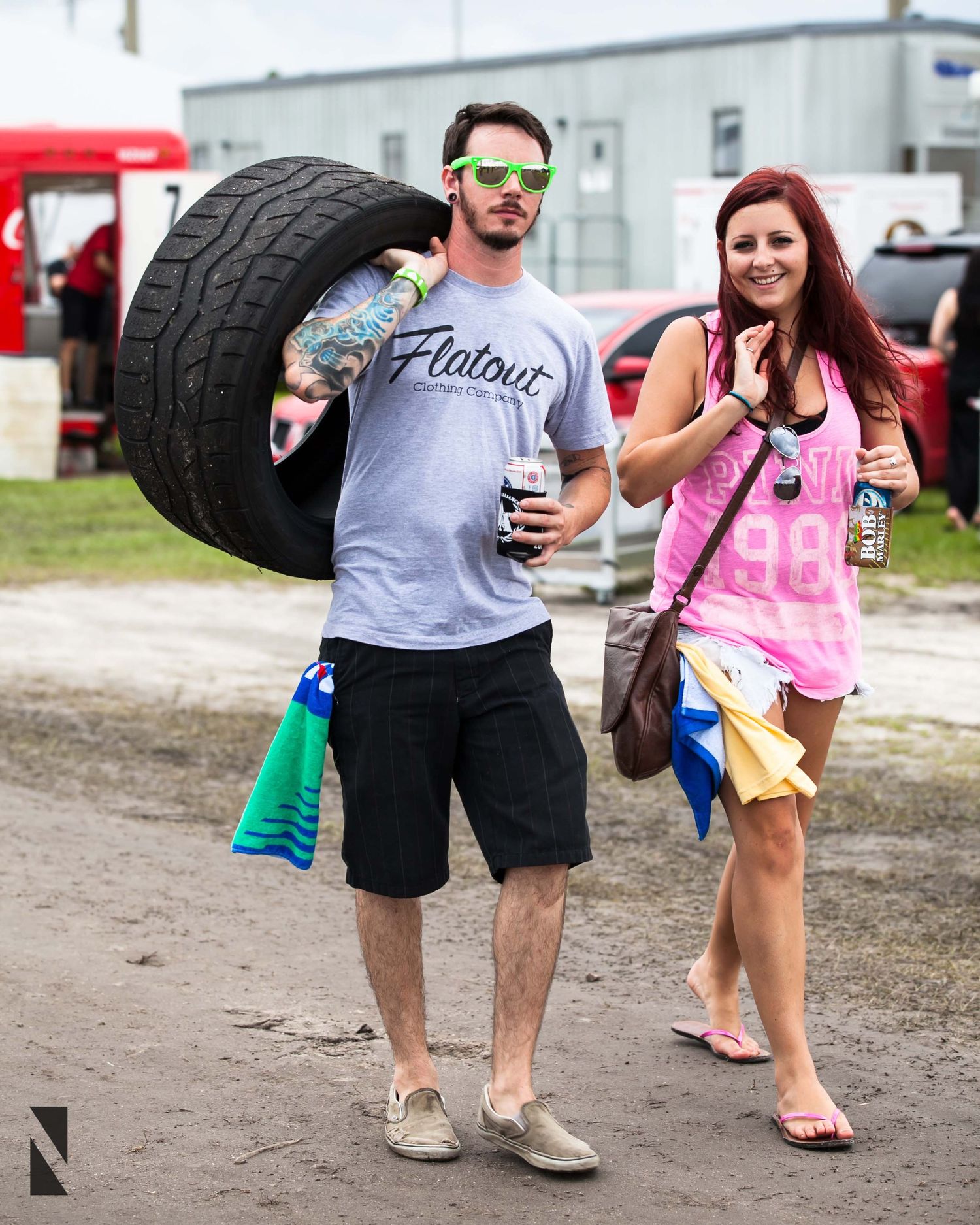 Festival attendees enjoy drinks outdoors, with one person carrying a large tire at an automotive event.