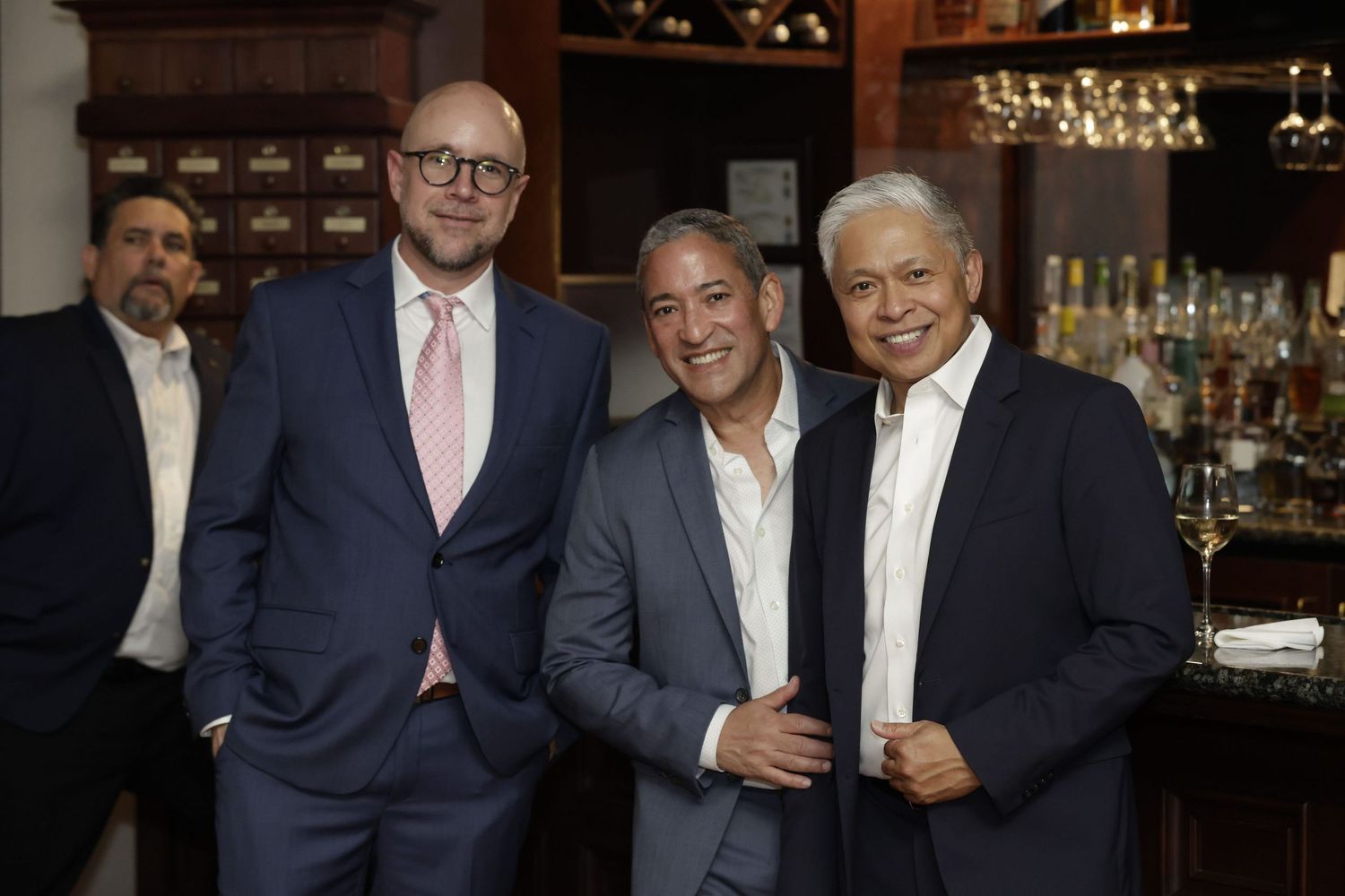 A group of men in formal attire stand together smiling at a bar with glasses and bottles in the background.