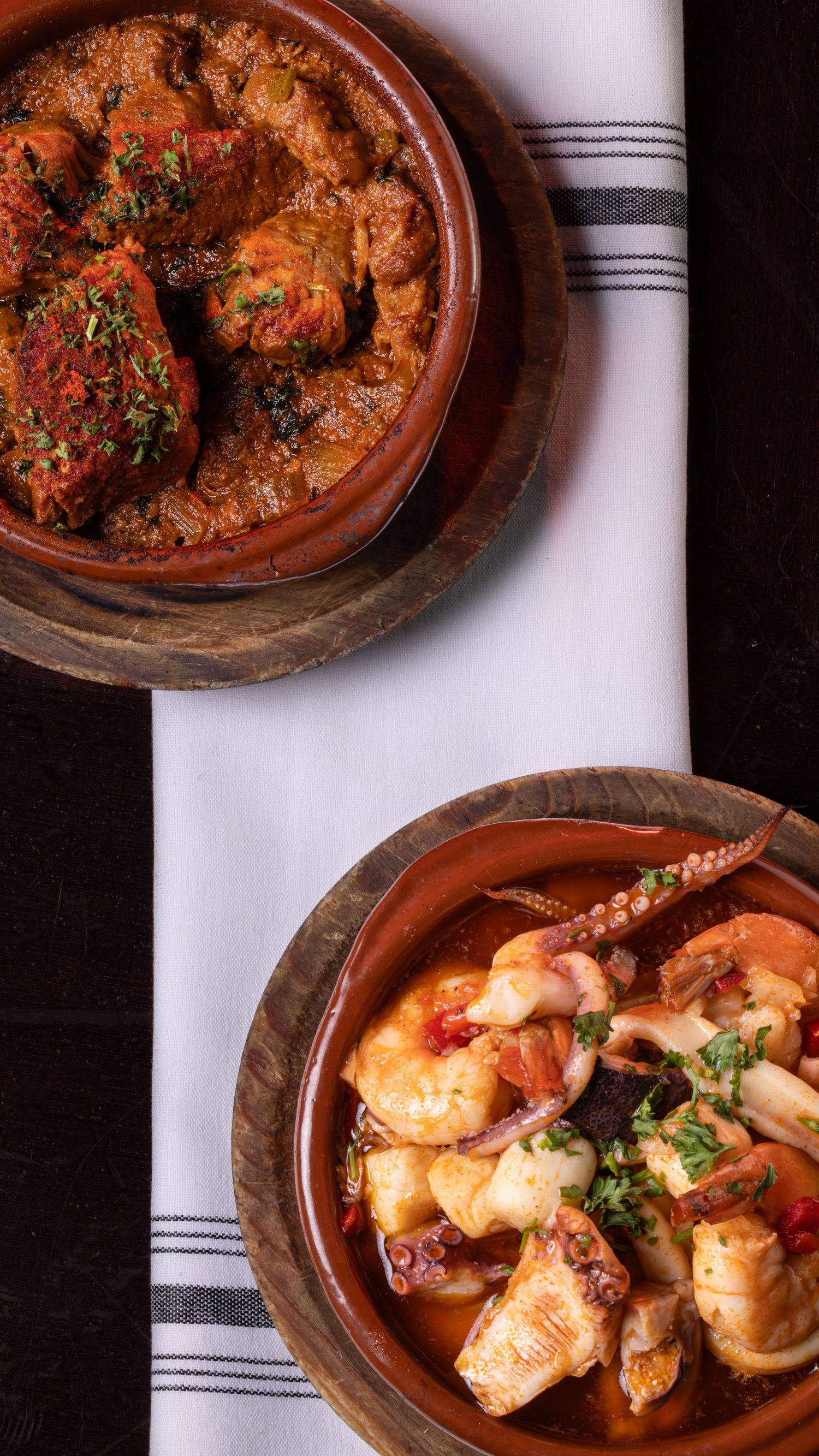 Close-up progression shots of shrimp and fish being cooked in a spicy reddish sauce in rustic clay serving dishes.