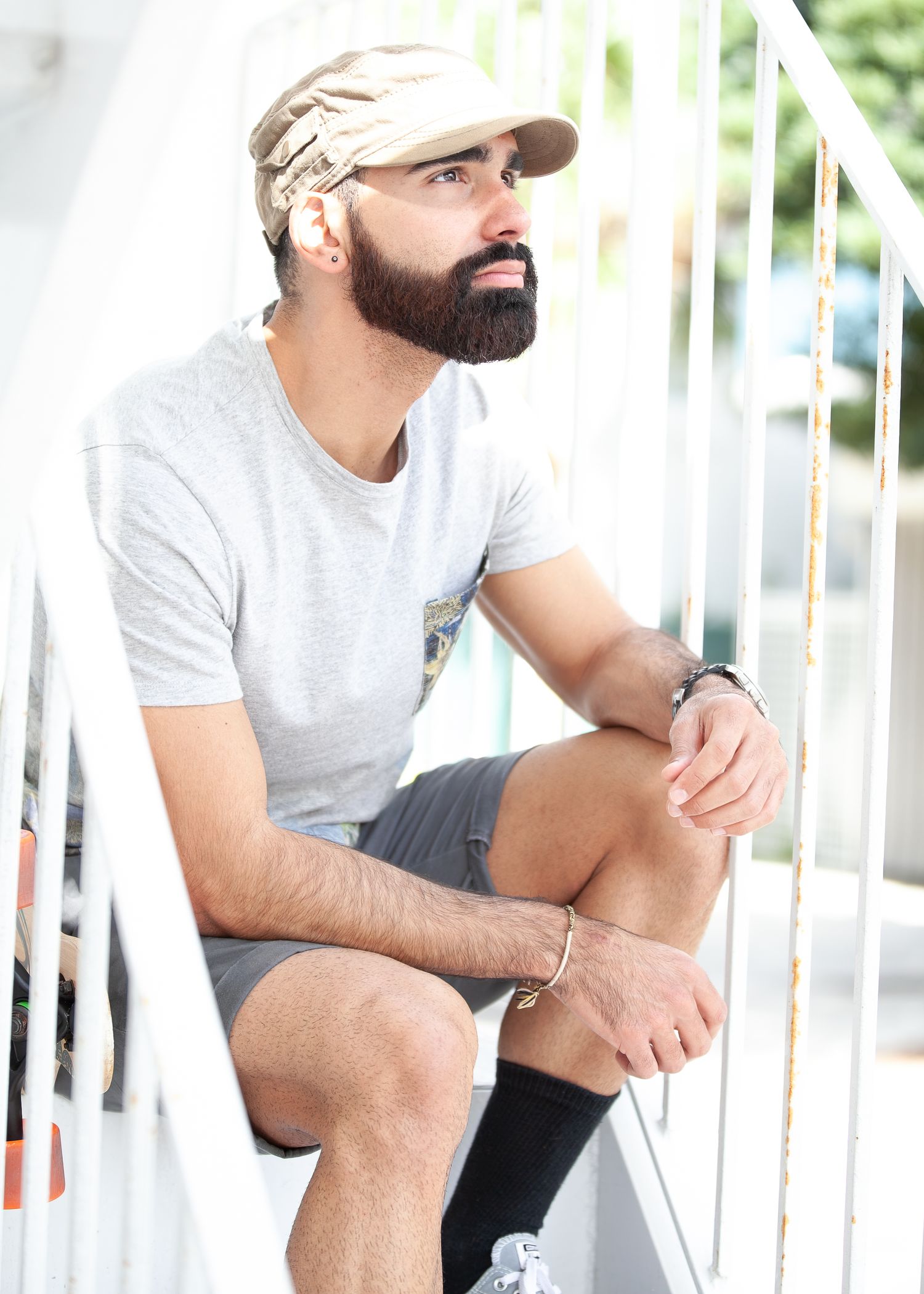 Person in a gray t-shirt and baseball cap sits thoughtfully on white stairs wearing shorts and dark socks.