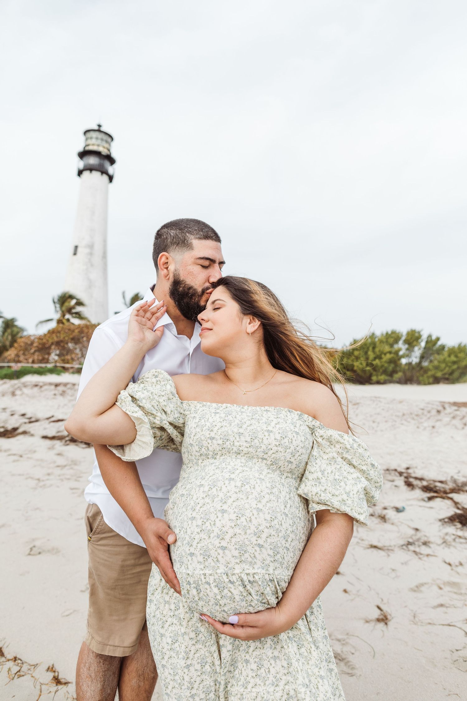 A romantic maternity photo shoot at a lighthouse beach with a couple embracing in front of the white tower structure.