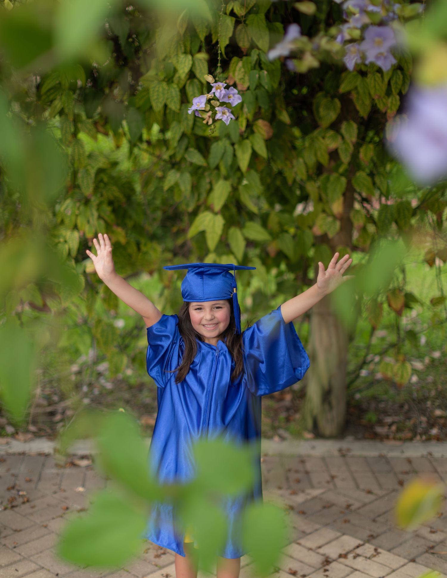 A graduate in bright blue cap and gown stands with arms raised joyfully on a brick pathway surrounded by green foliage.