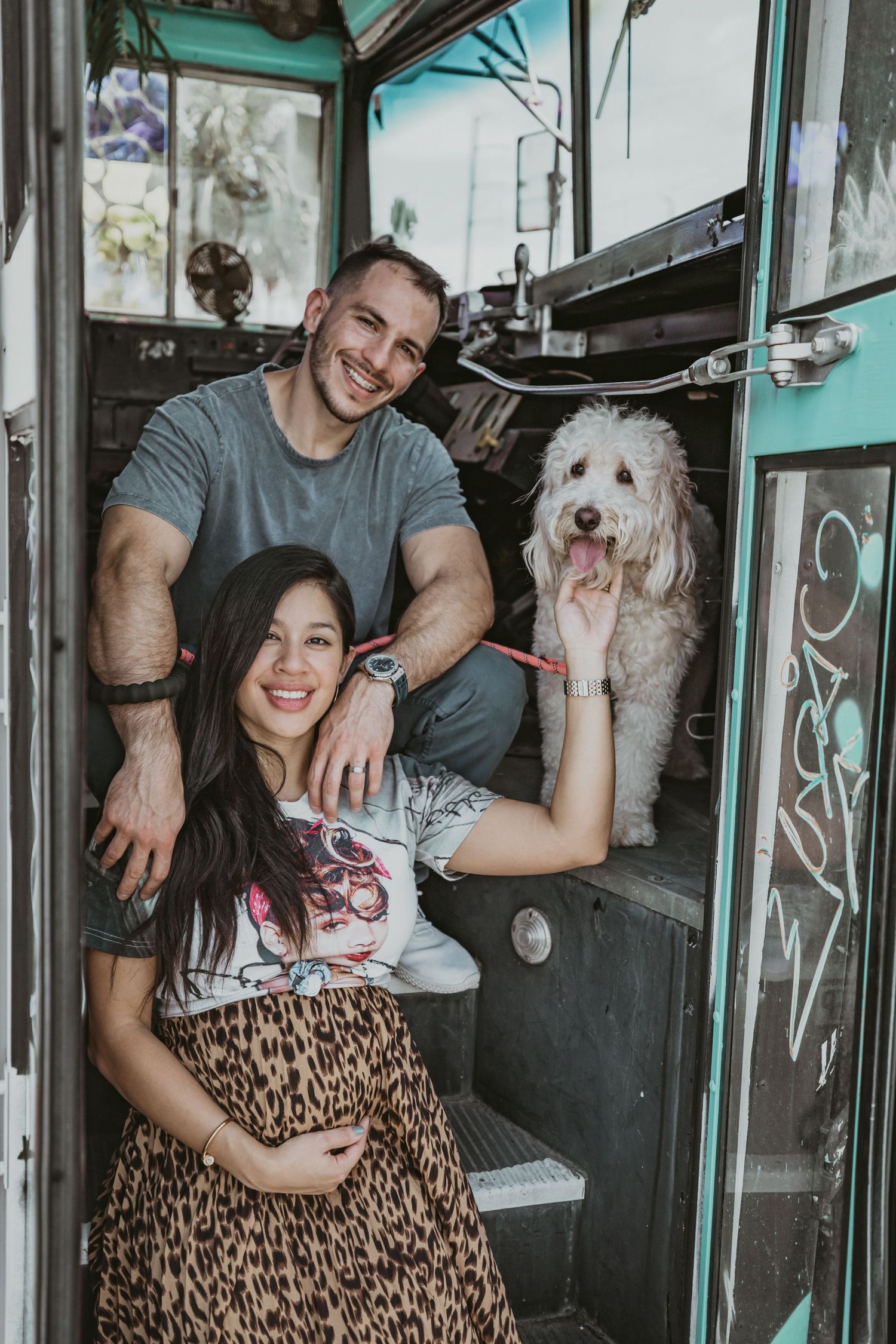 A couple poses playfully with their white fluffy dog inside what appears to be a vintage vehicle.