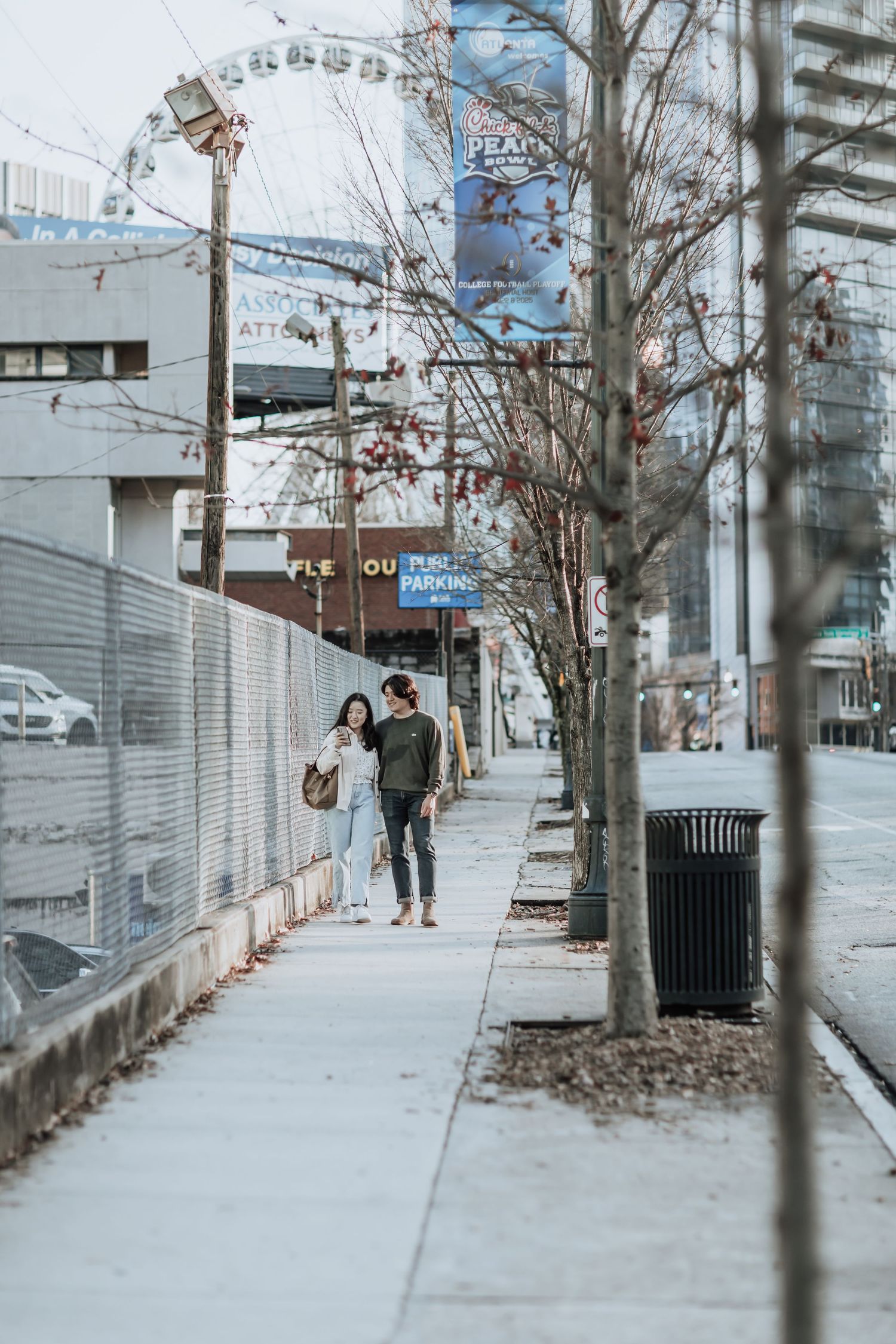 A couple strolls down an urban sidewalk with tall buildings and a ferris wheel visible in the background.
