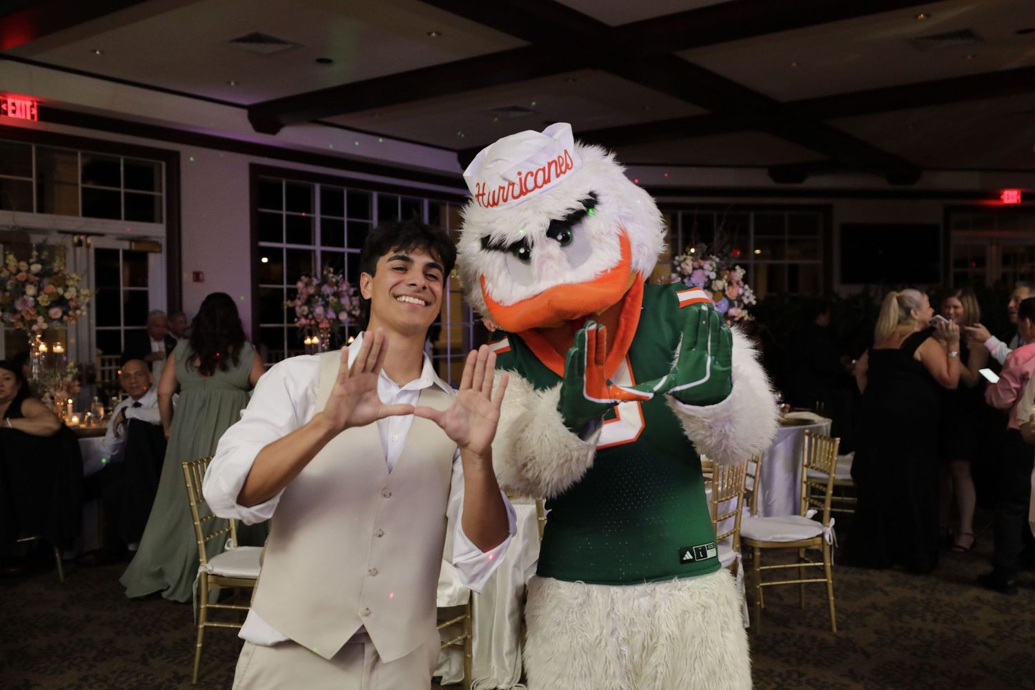 Someone in a beige vest poses with a sports mascot wearing a green Miami Hurricanes jersey at an indoor event.
