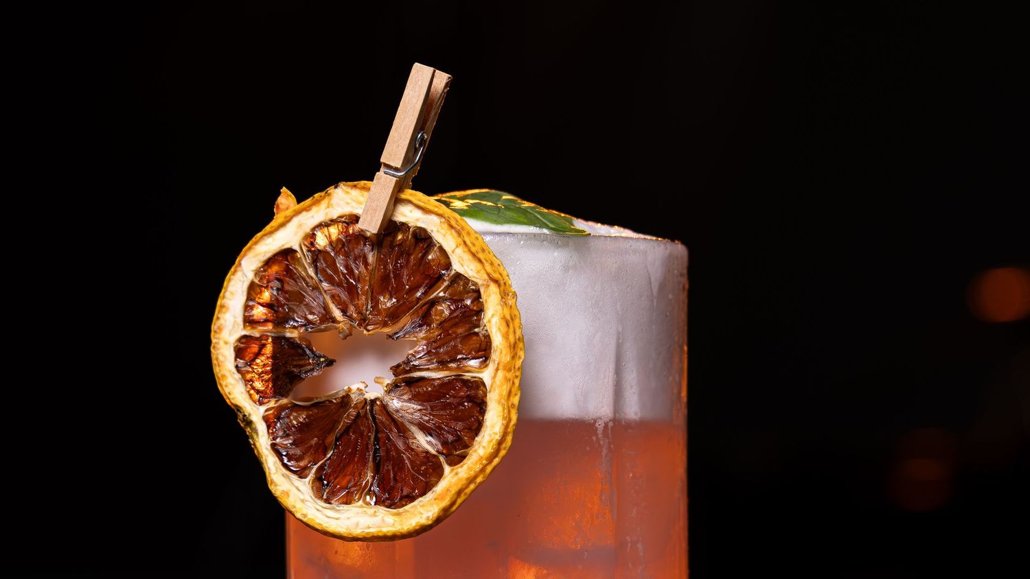 Iced cocktail garnished with a dried orange slice on a black background with a copper straw.
