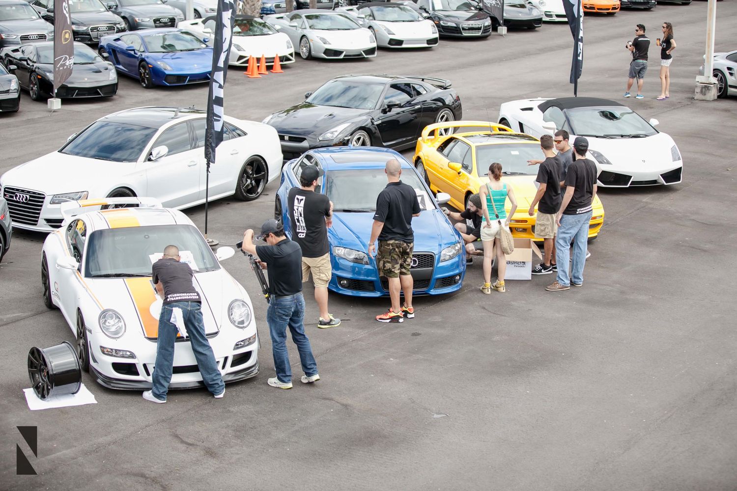 A group of people gathered around luxury sports cars including Porsche and Lamborghini models in a parking lot.