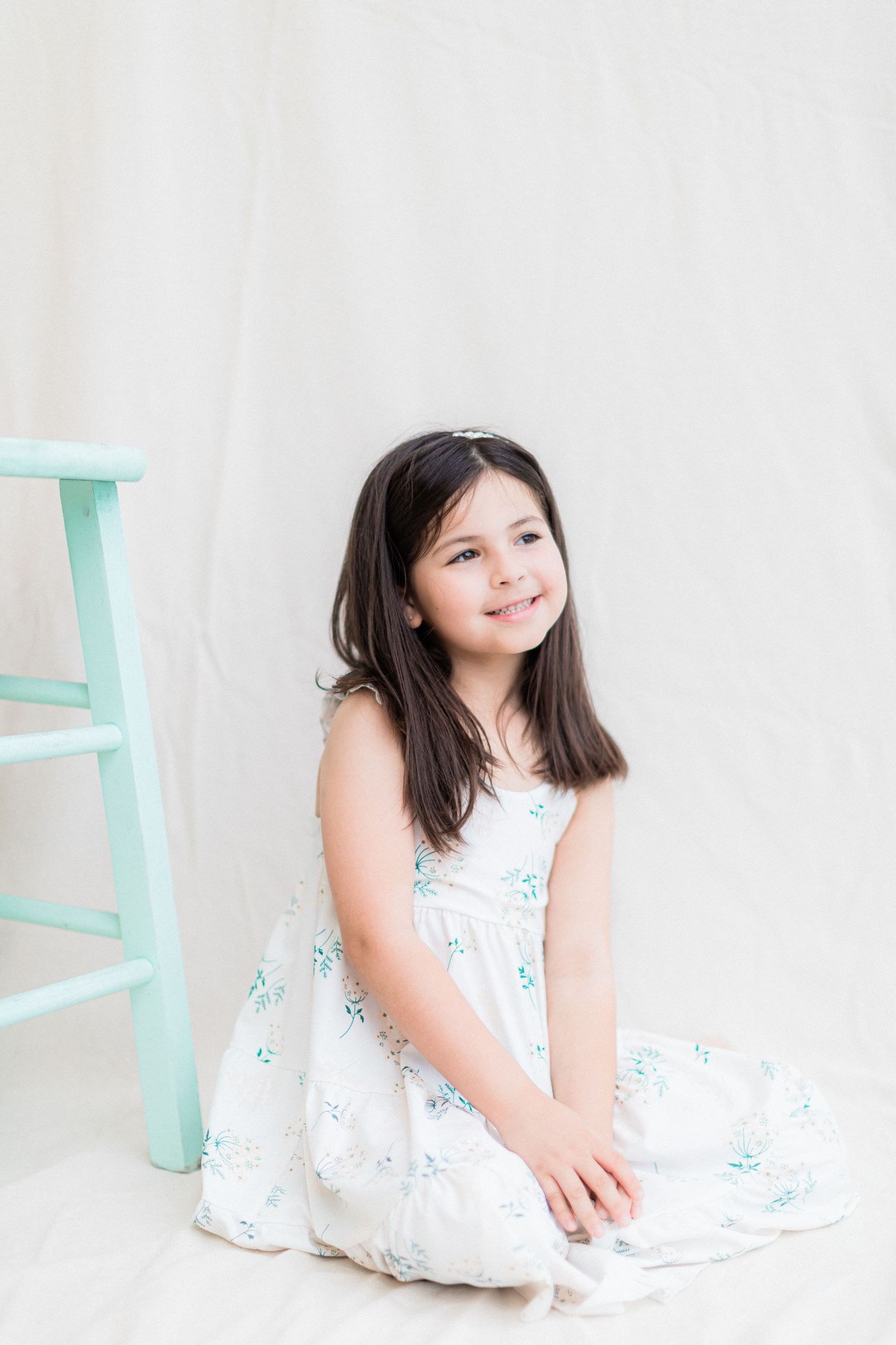 A young girl in a white floral dress sits sweetly against a light background with a mint green stool.