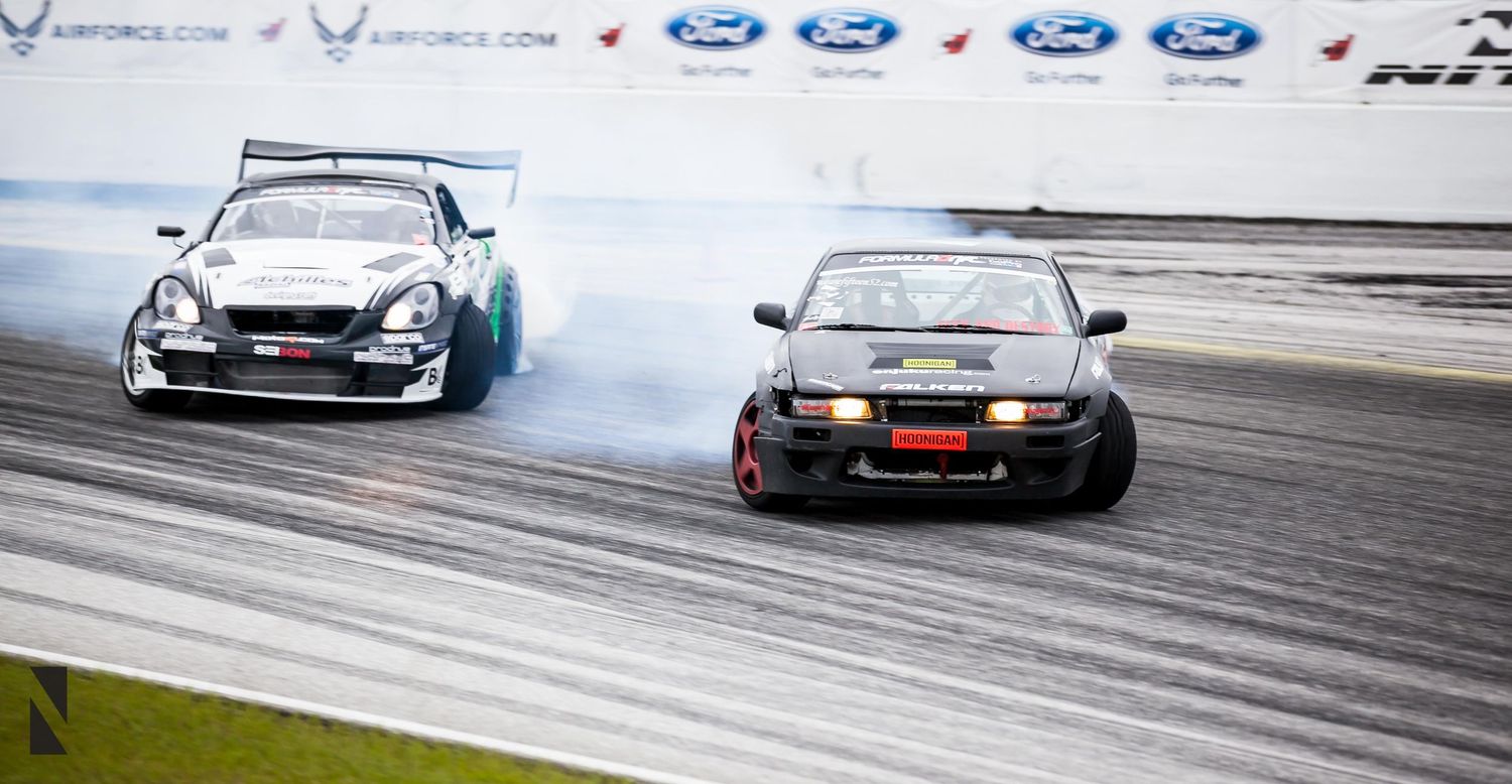 Two drift cars sliding in tandem through a corner leaving tire smoke trails on the track.