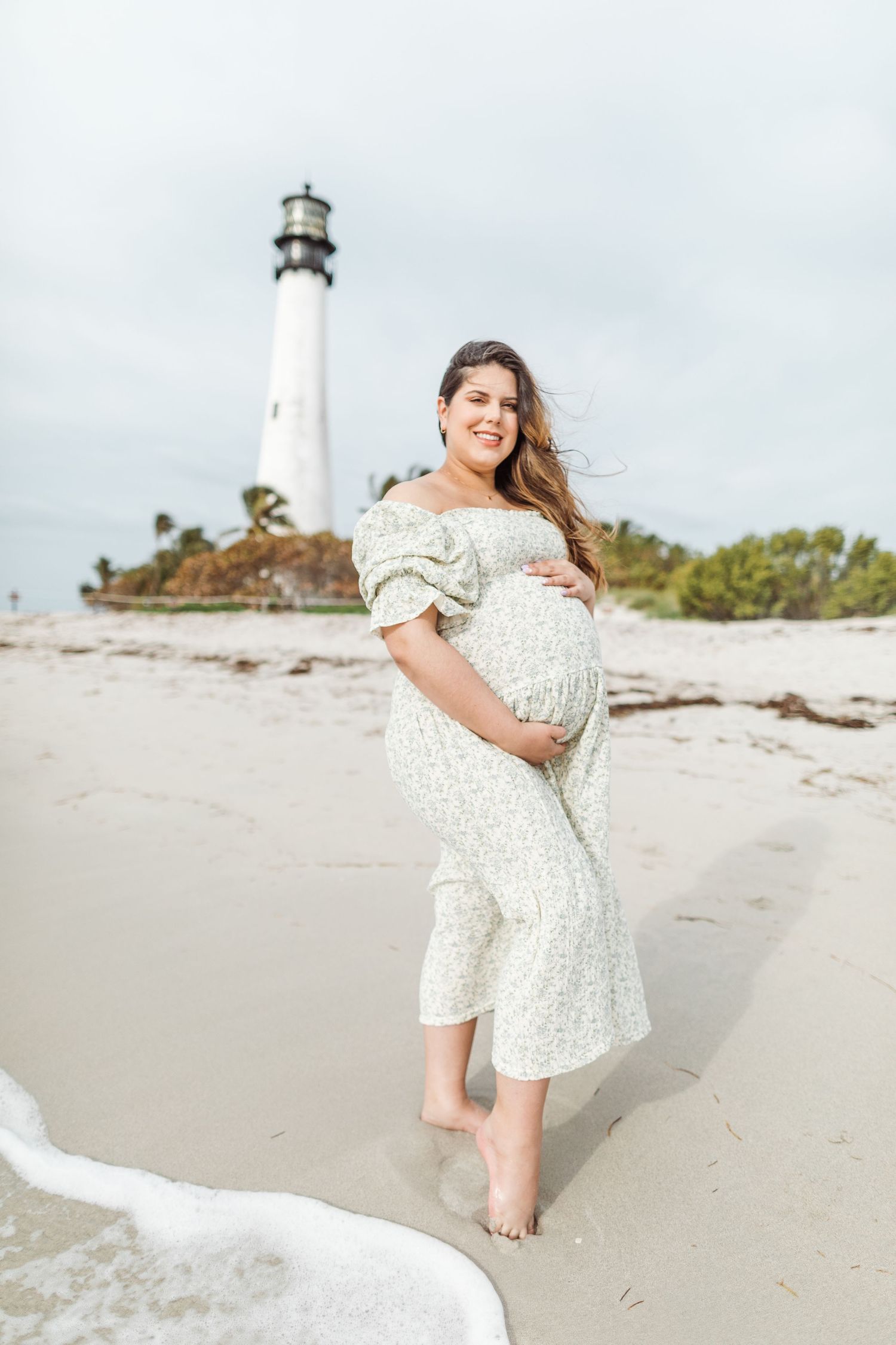 A pregnant woman in a white floral dress poses barefoot on a beach with a lighthouse in the background.