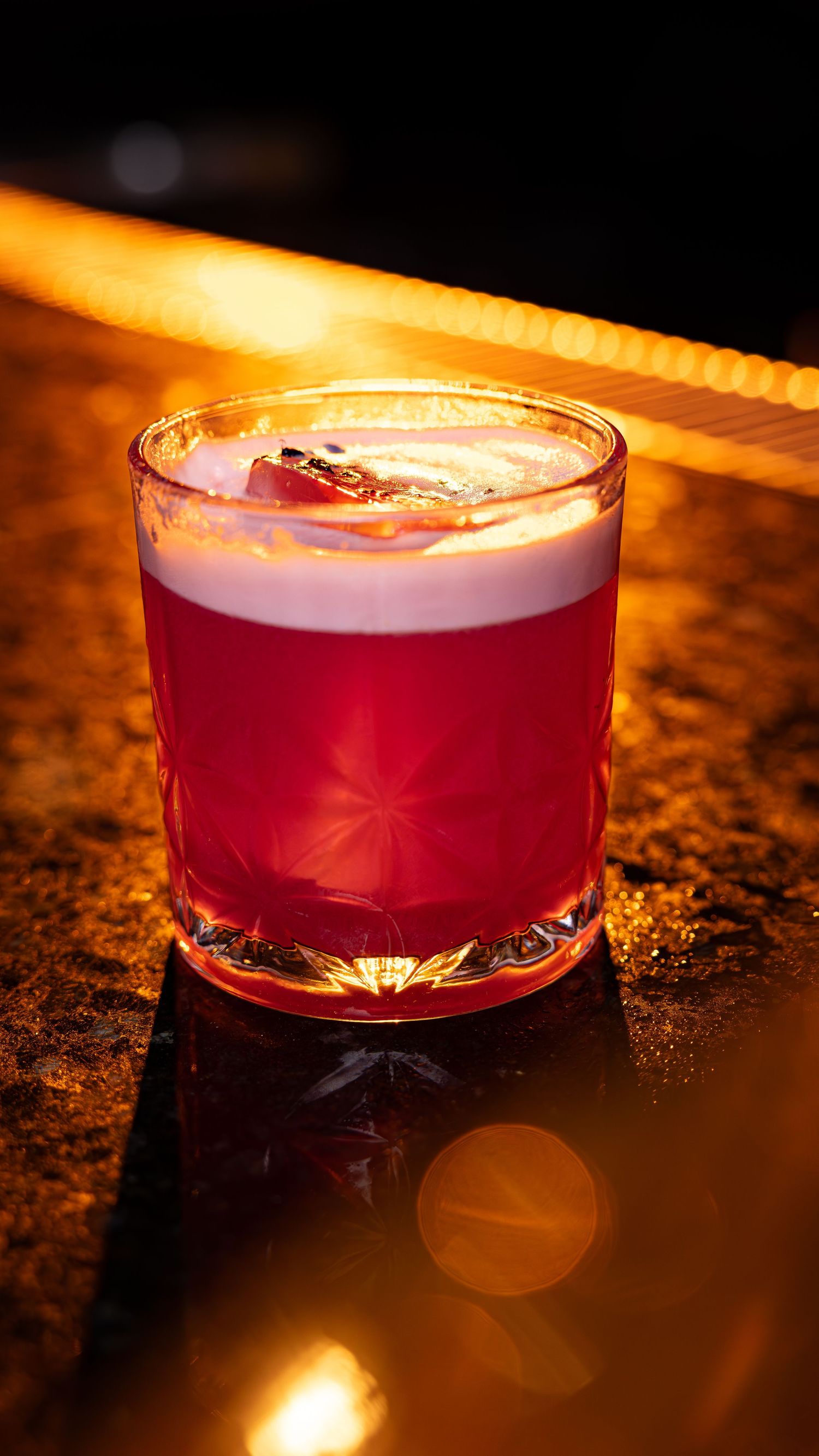 A vibrant red cocktail with foam top served in a rocks glass on a dark surface with warm ambient lighting.