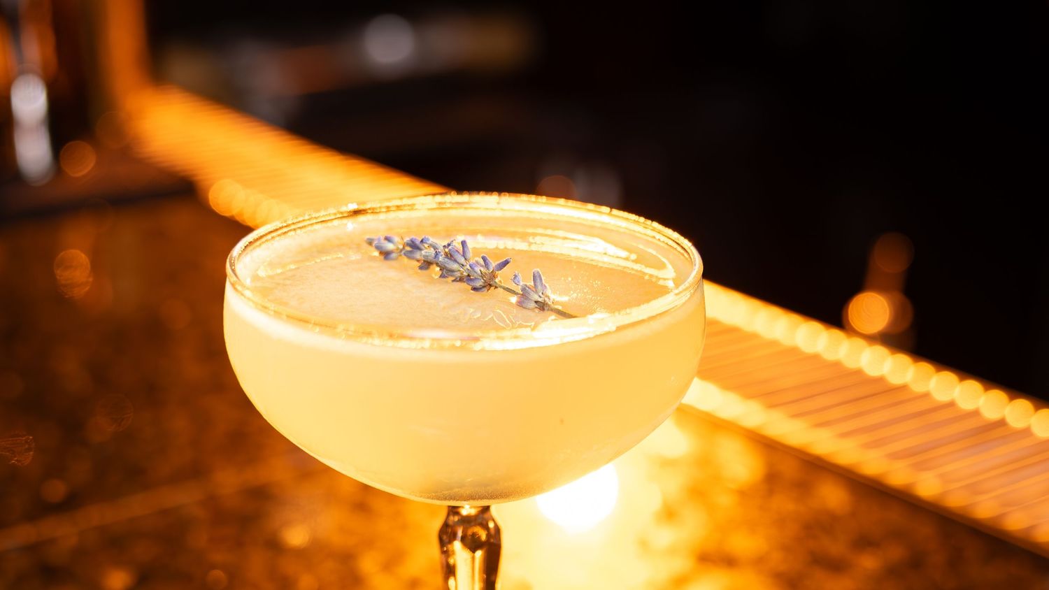A golden cocktail garnished with lavender buds served in a coupe glass on a dimly lit bar counter.