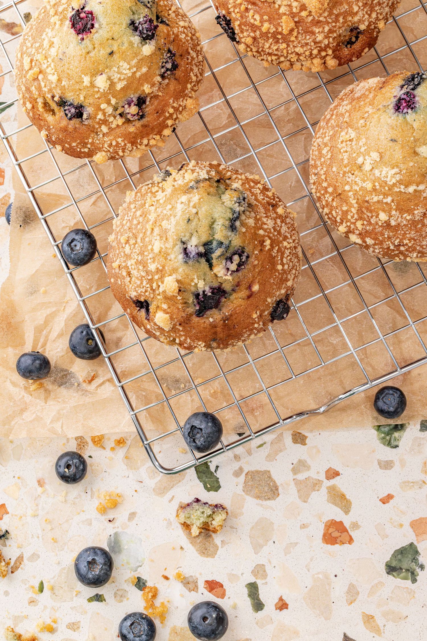 Freshly baked blueberry muffins with golden streusel topping cooling on a wire rack with scattered berries and crumbs.