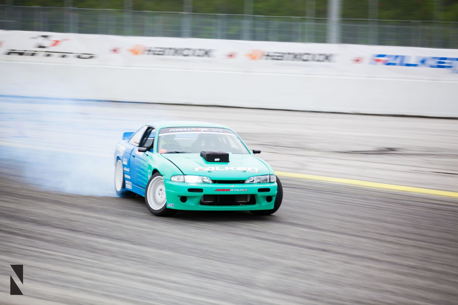 A turquoise drift car creates smoke while sliding sideways on a racetrack during a drifting competition.