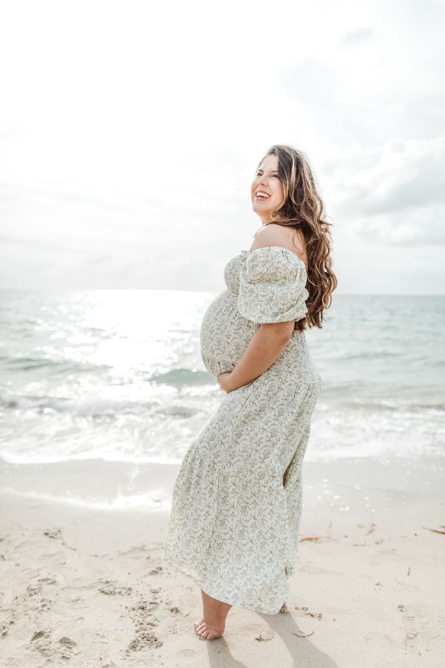 Expectant woman in flowy floral dress smiles while standing barefoot on sunny beach with ocean waves in background.