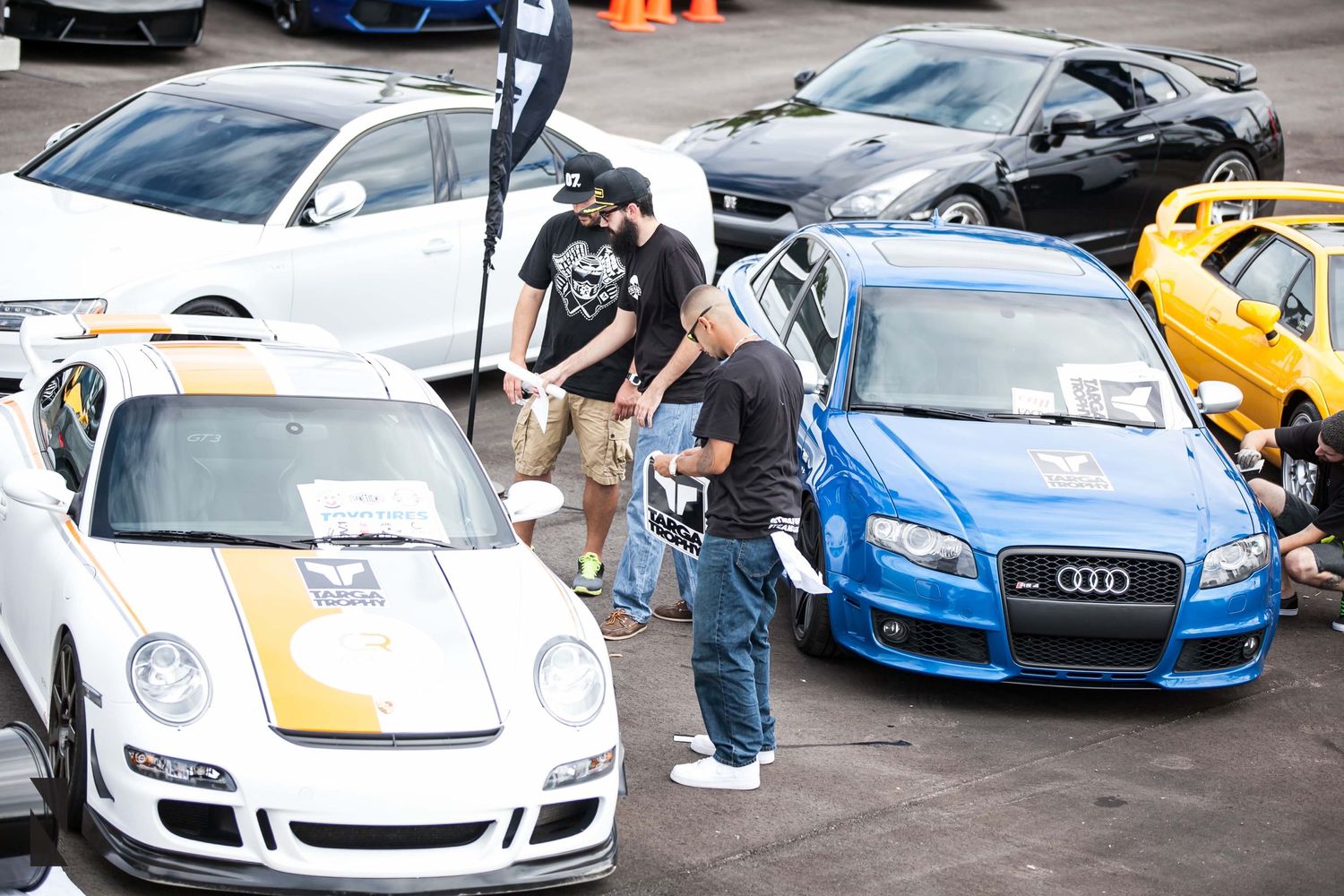 Car enthusiasts examining custom vehicles including a Porsche and Audi at an outdoor car show.