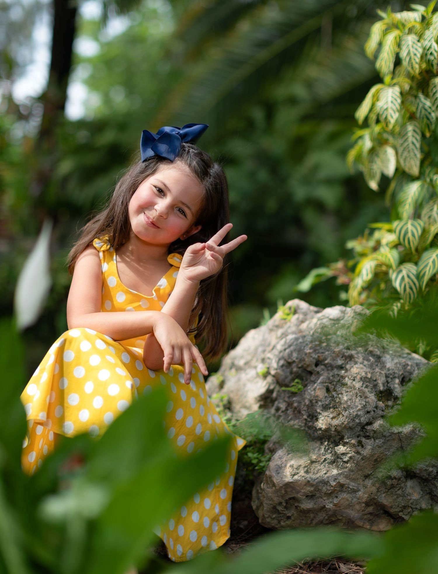 A cheerful figure in a yellow polka dot dress poses on rocks surrounded by green foliage on a sunny day.