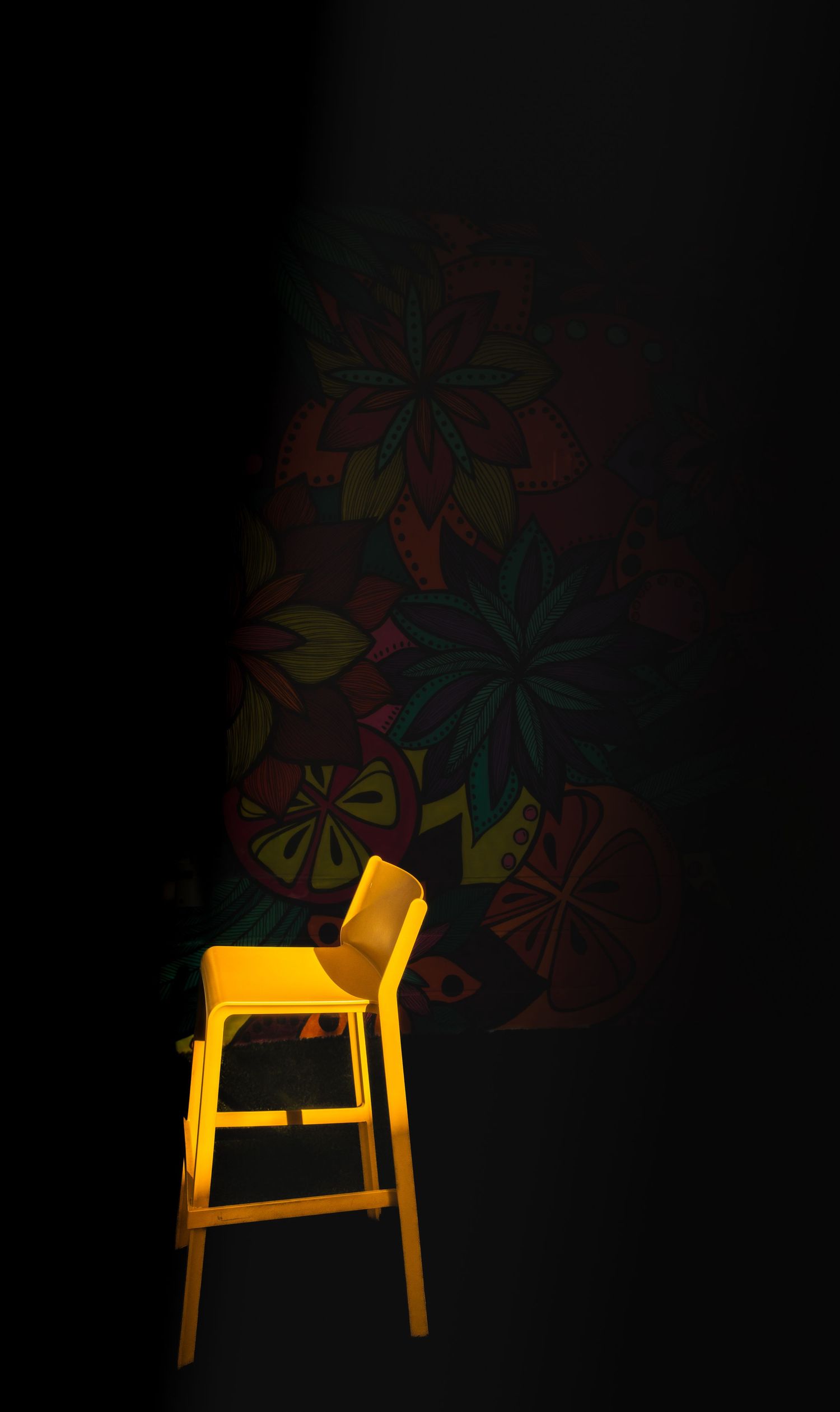 Yellow barstool against dark wall with colorful floral pattern shadow.