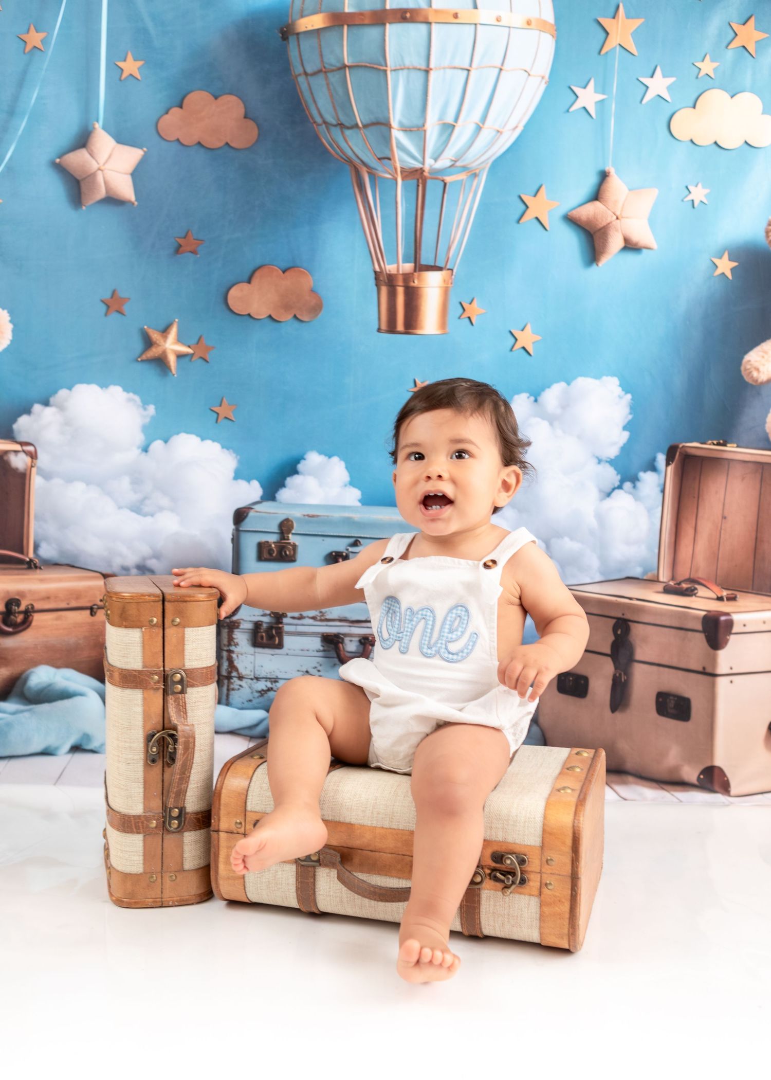 Joyful baby sits on antique suitcase with dreamy sky background and floating stars.