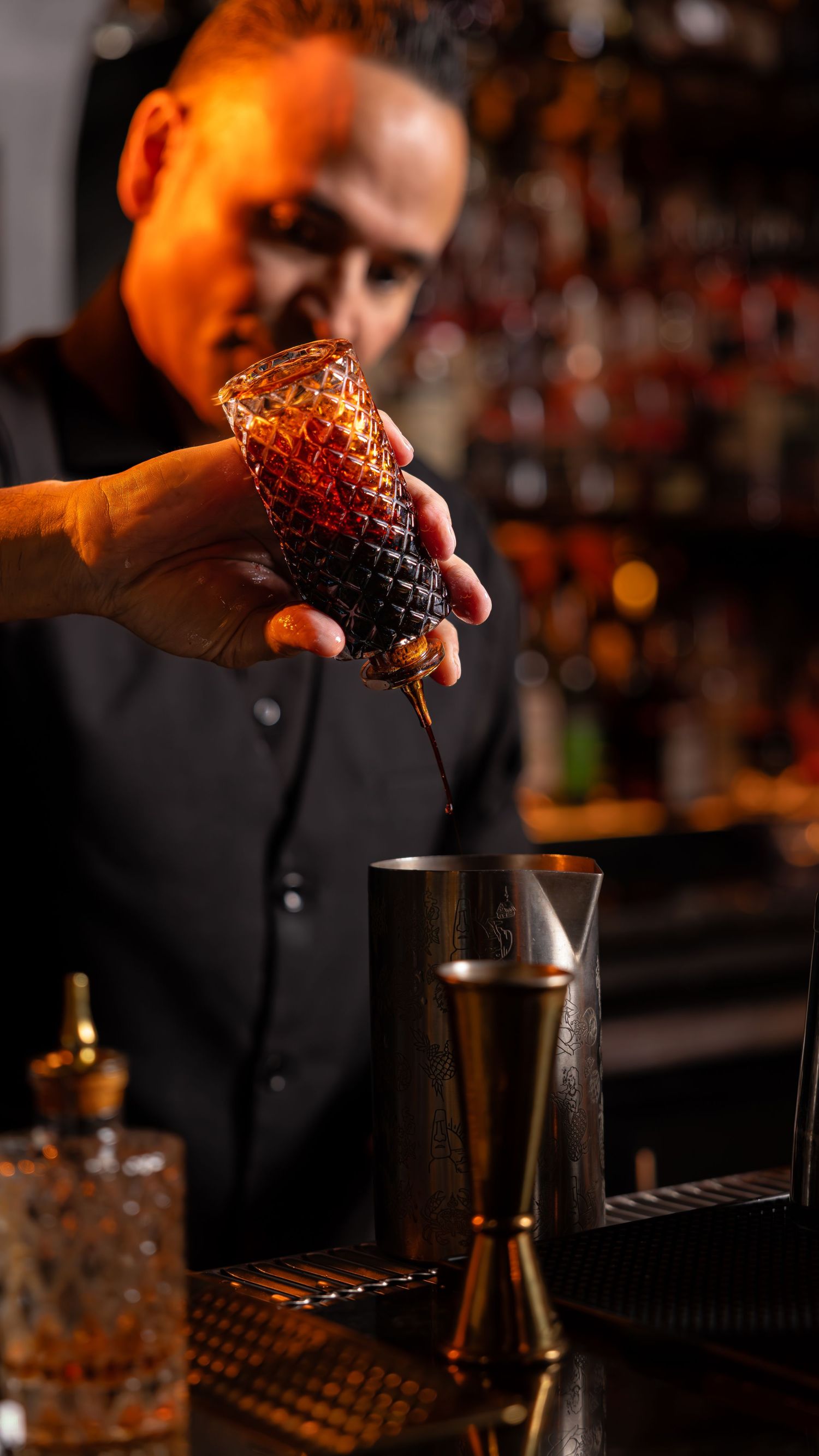 A bartender in black attire carefully pours a cocktail using an ornate glass container in a dimly lit bar setting.