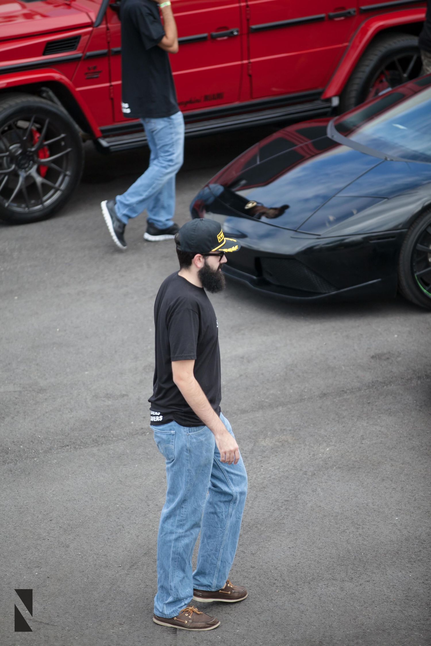 Person in black t-shirt and jeans stands in parking lot next to luxury vehicles including a red G-Wagon and dark supercar.