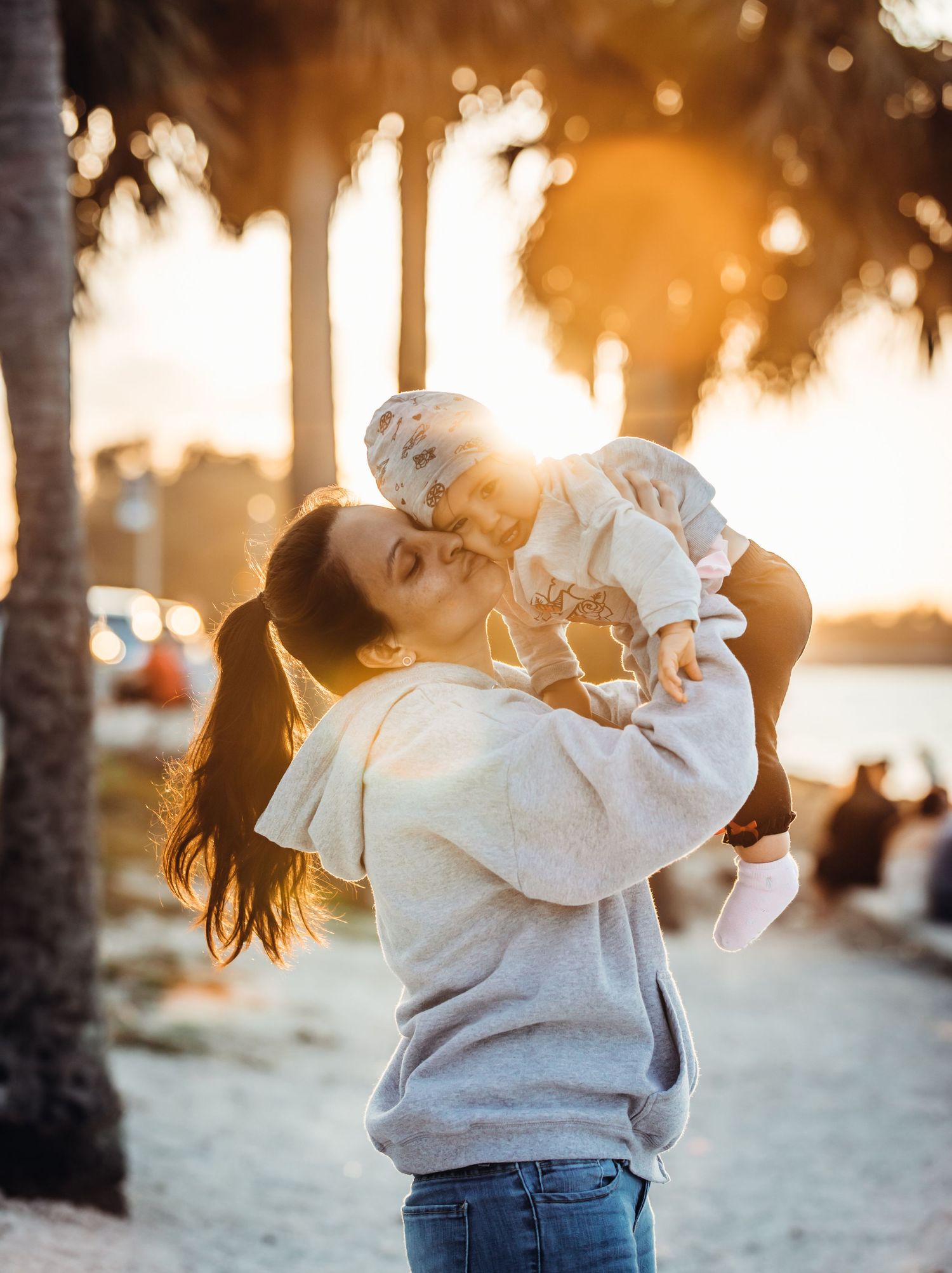Parent lifting and kissing a baby against a golden sunset backdrop with palm trees at the beach.