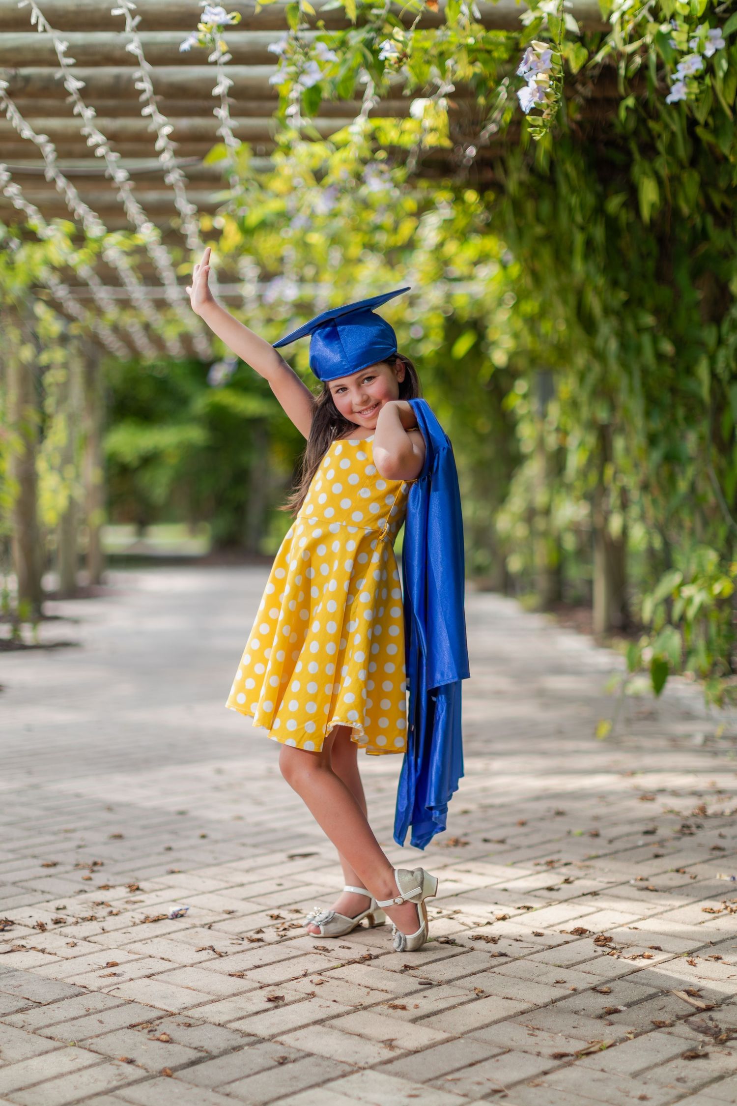 A blue graduation cap and gown with a bright yellow polka dot dress twirls in a garden pathway setting.