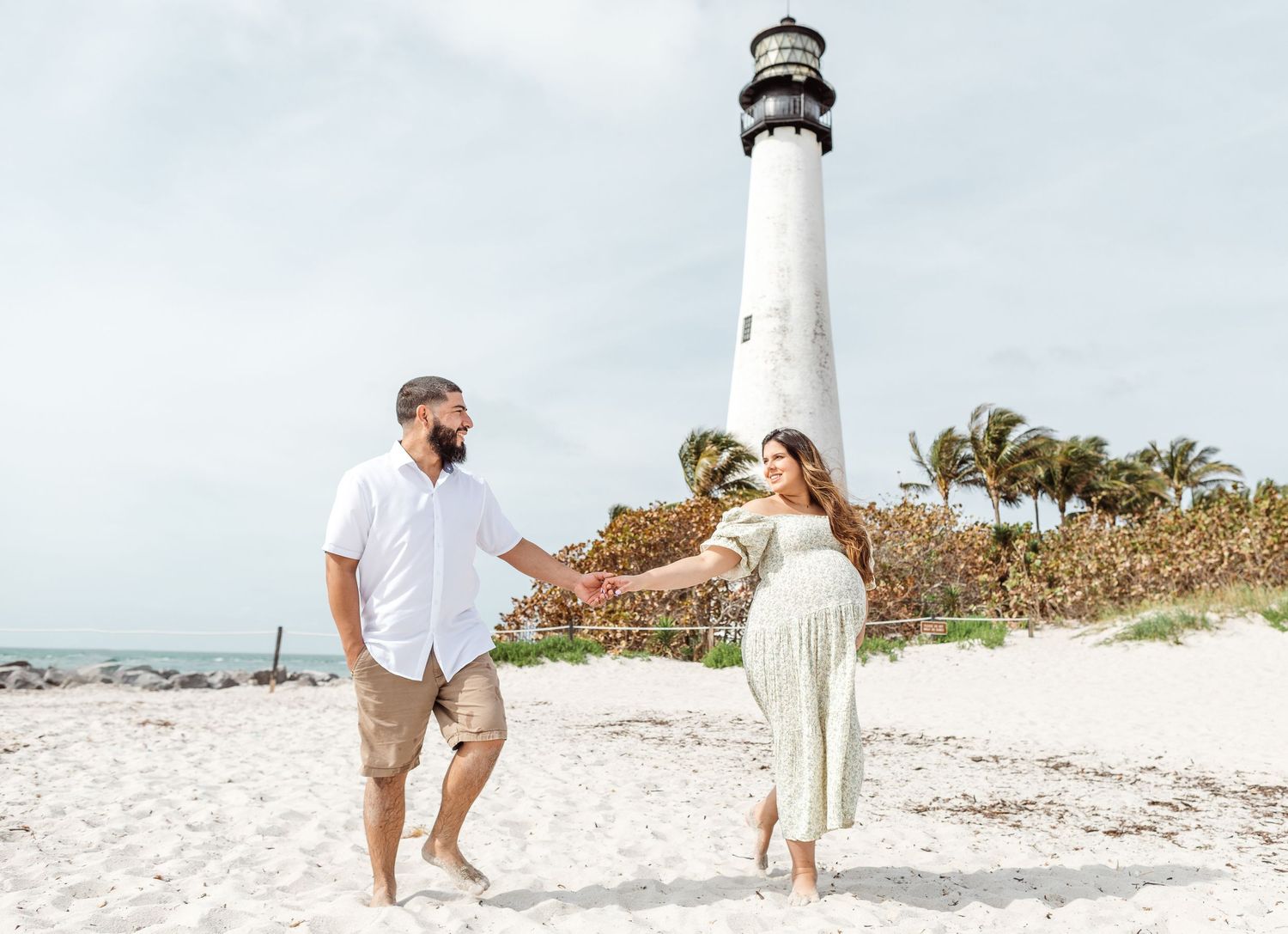 Two people walking on sandy beach with lighthouse and palm trees in background on overcast day.