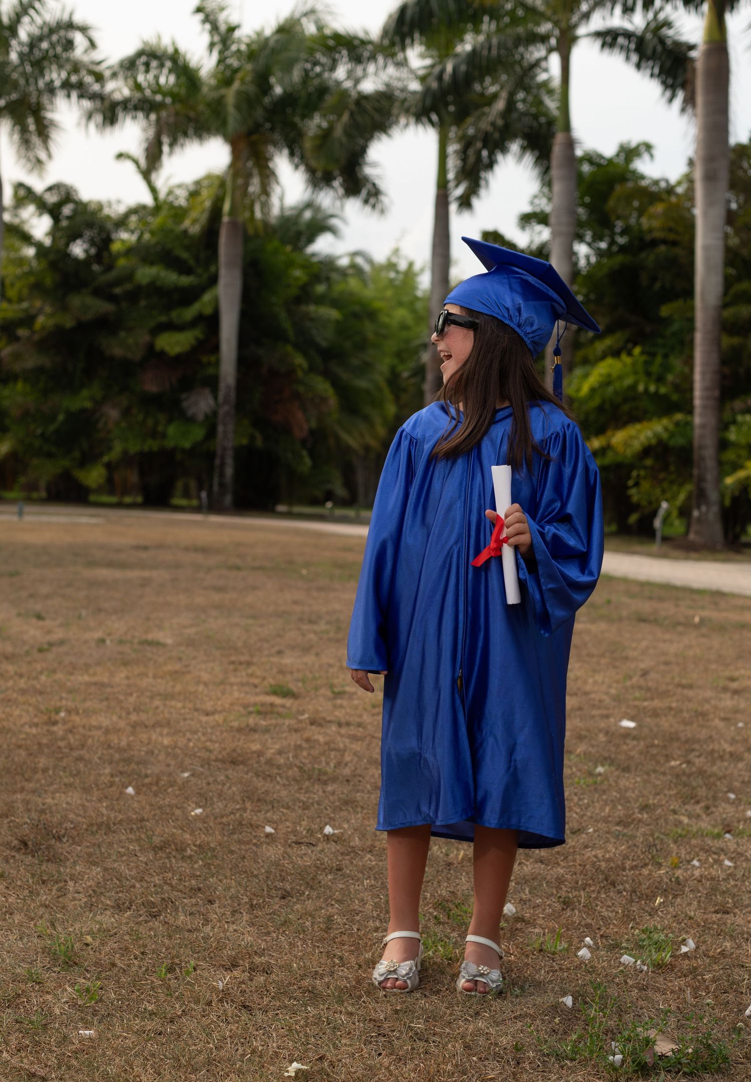 A graduate in a royal blue cap and gown stands outdoors in front of palm trees while holding a diploma.