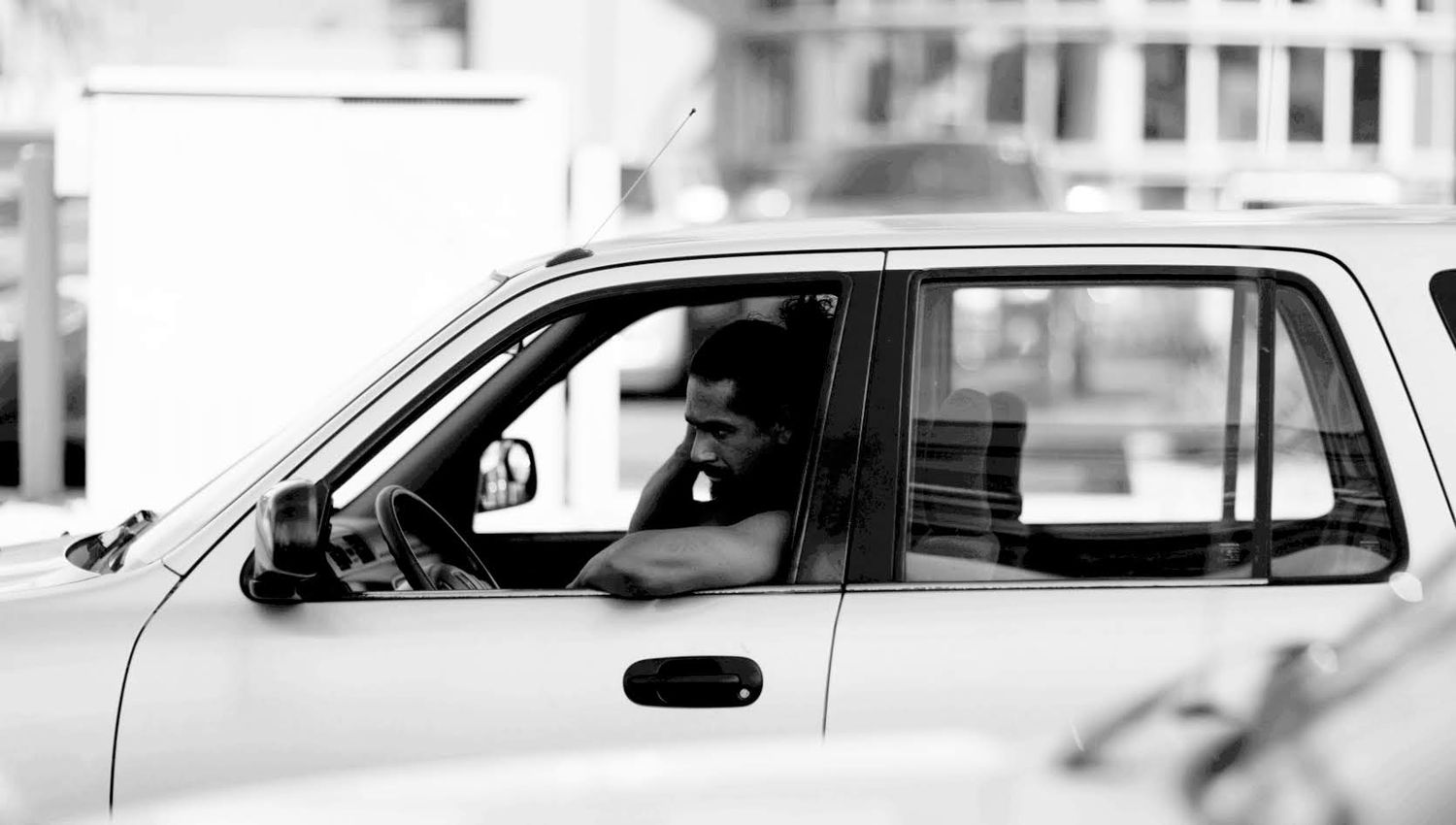 Black and white photo of someone driving a car viewed through the driver's side window.