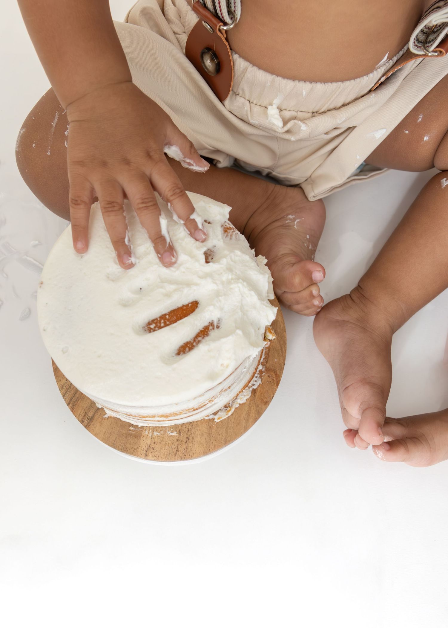 Small hands spreading white whipped cream or frosting on top of a round cake on a wooden cutting board in steps.
