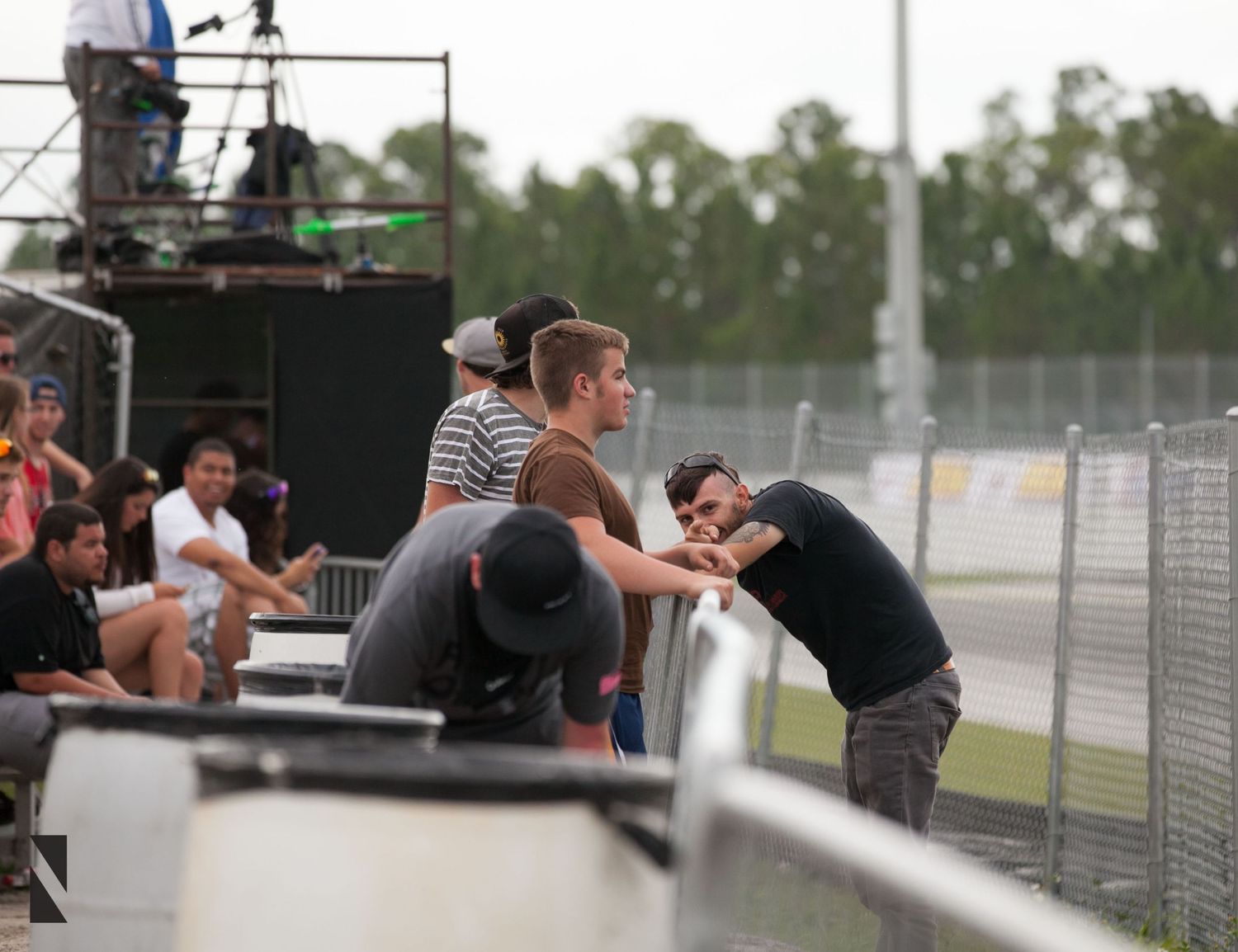 Race spectators lean against a fence barrier watching trackside activity during a motorsport event.