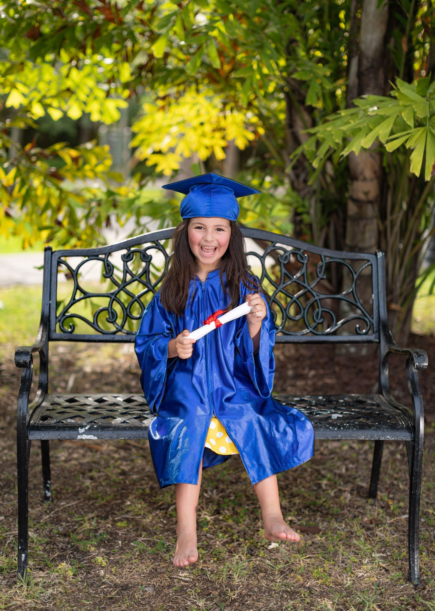 A graduate in bright blue cap and gown sits smiling on an ornate iron bench surrounded by lush greenery.