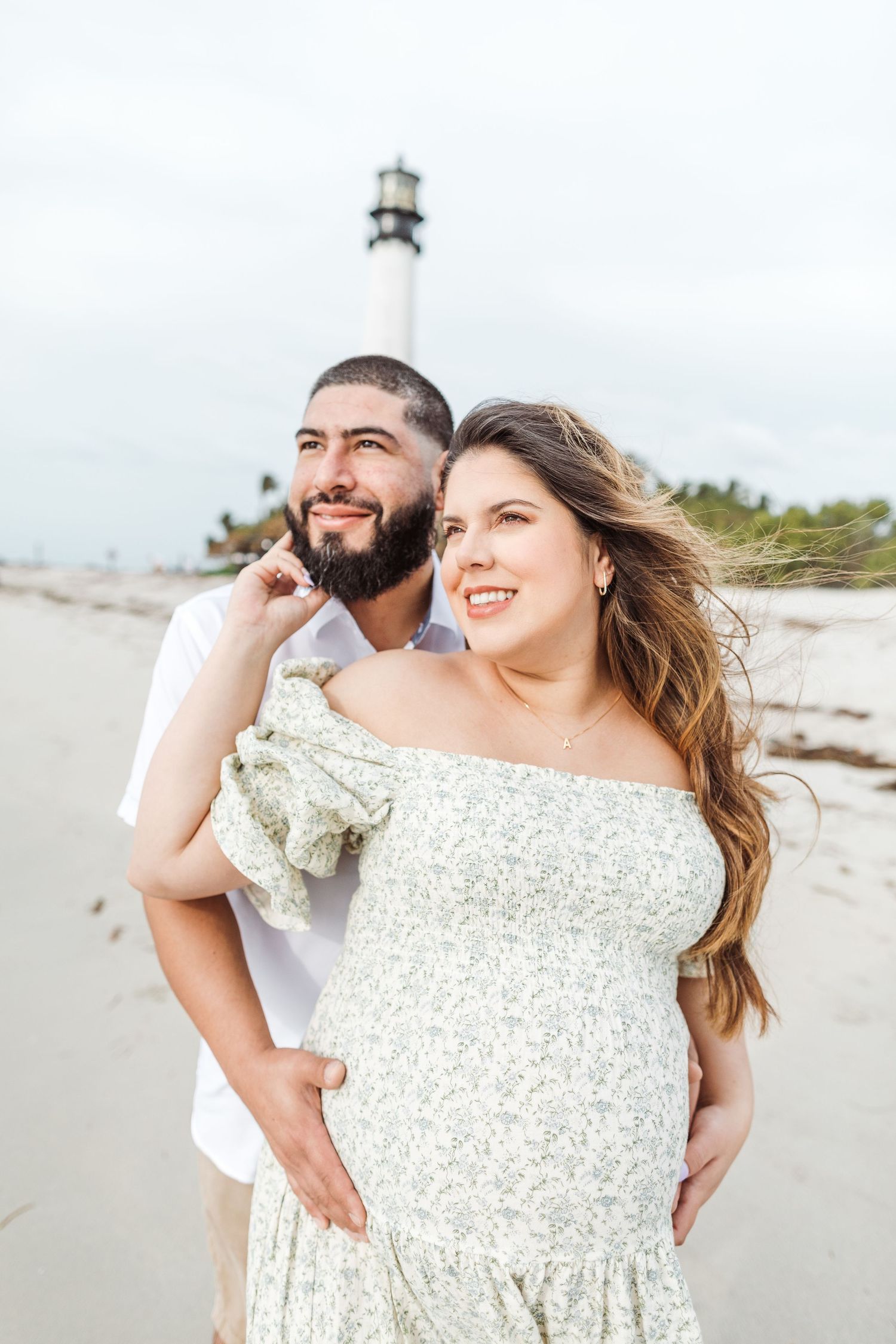A couple enjoys a romantic moment on a beach with a lighthouse visible in the background on a cloudy day.