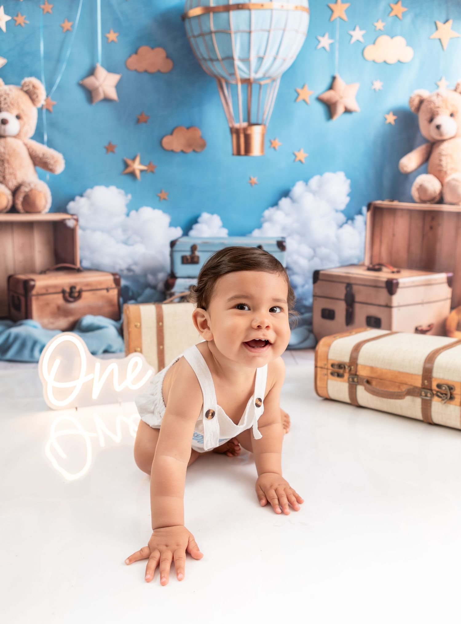 Adorable baby in white outfit crawls on floor with vintage suitcases, teddy bears, and hot air balloon backdrop.