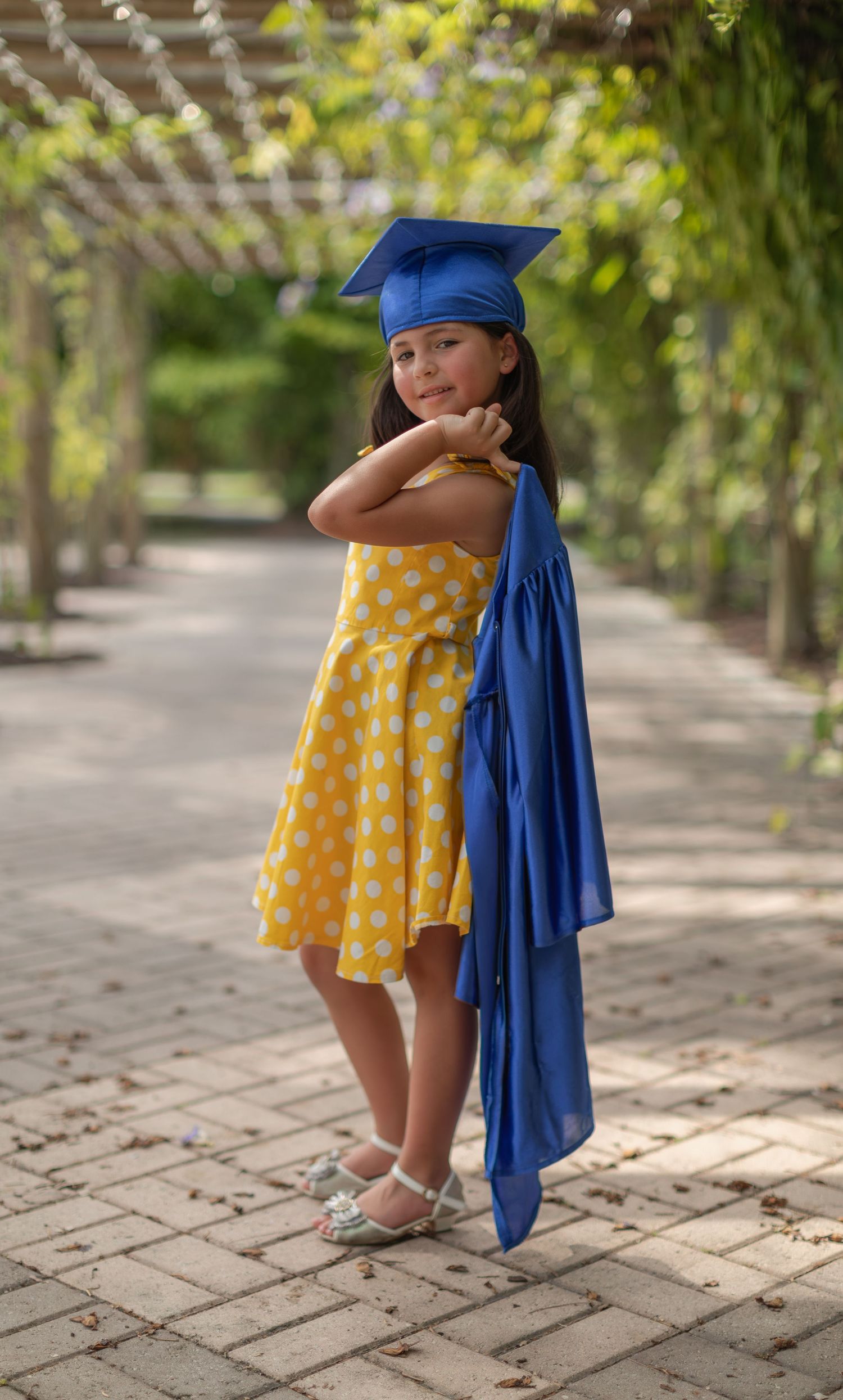 A young graduate in a blue cap and gown poses playfully wearing a yellow polka dot dress in an outdoor garden setting.