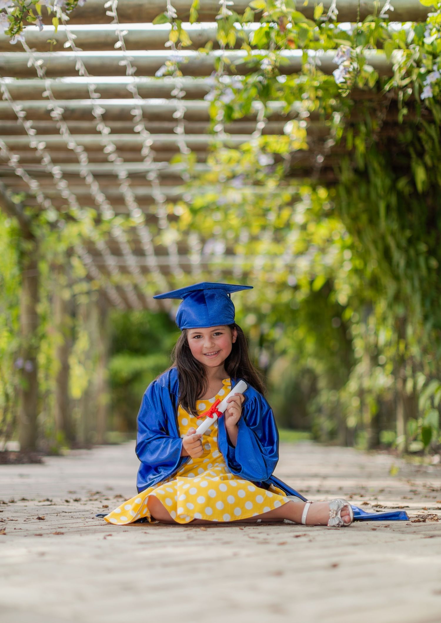 A graduate in blue cap and gown sits in a garden tunnel with trailing vines and a polka dot yellow dress.