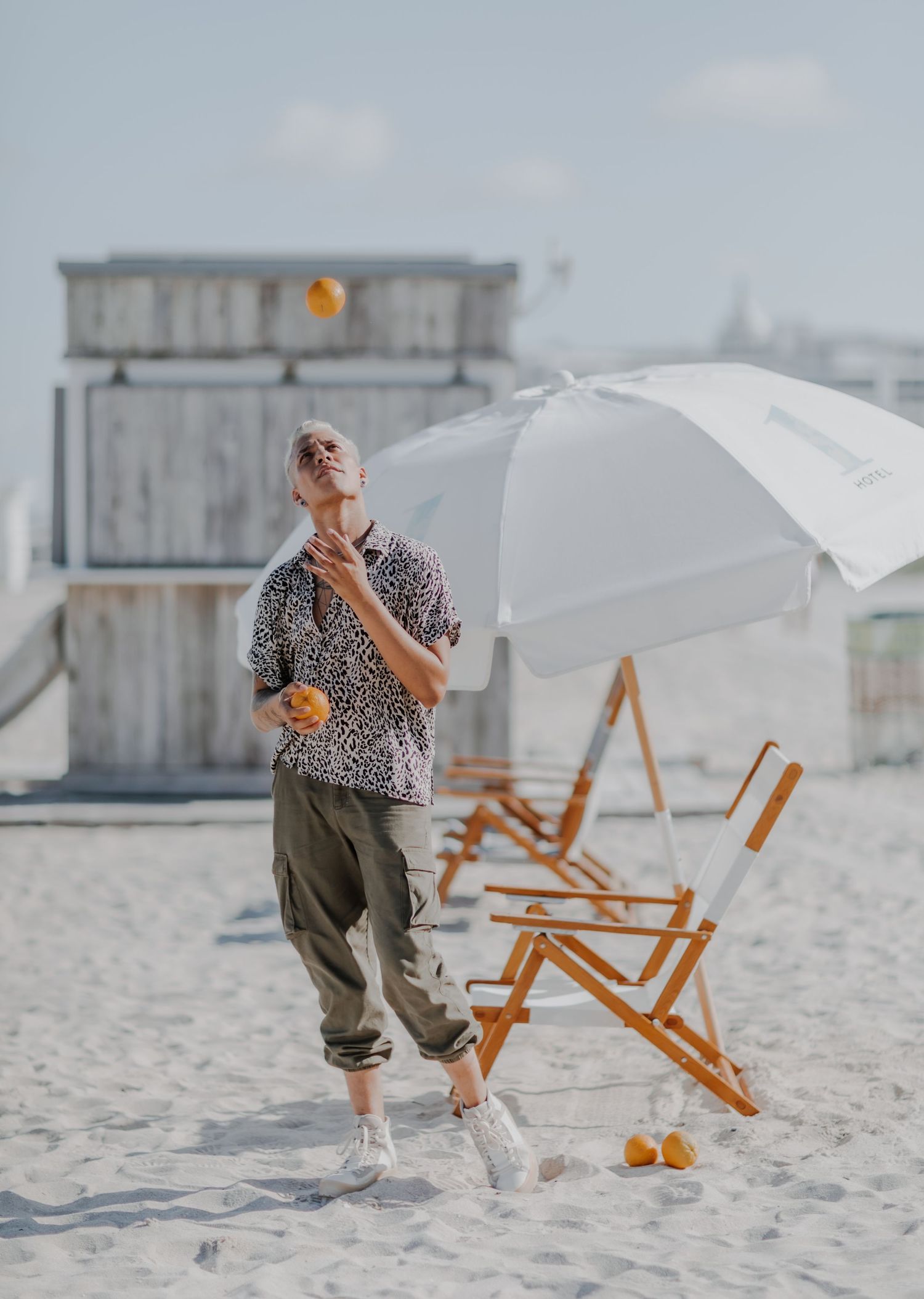 Person in animal print shirt juggling oranges on sunny beach with beach chair.