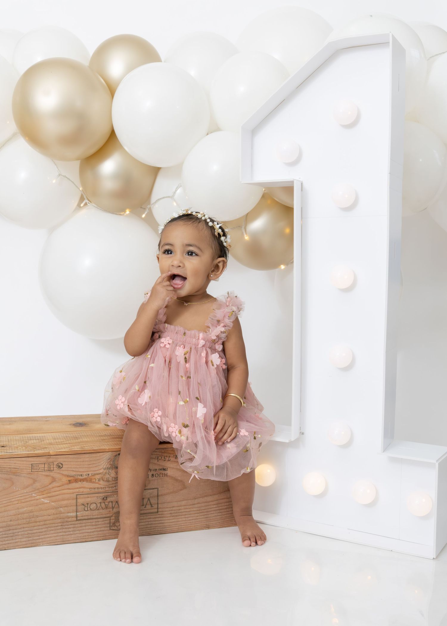 One year old in pink ruffle dress sits on rustic wood floor surrounded by gold and white balloons for birthday photos.