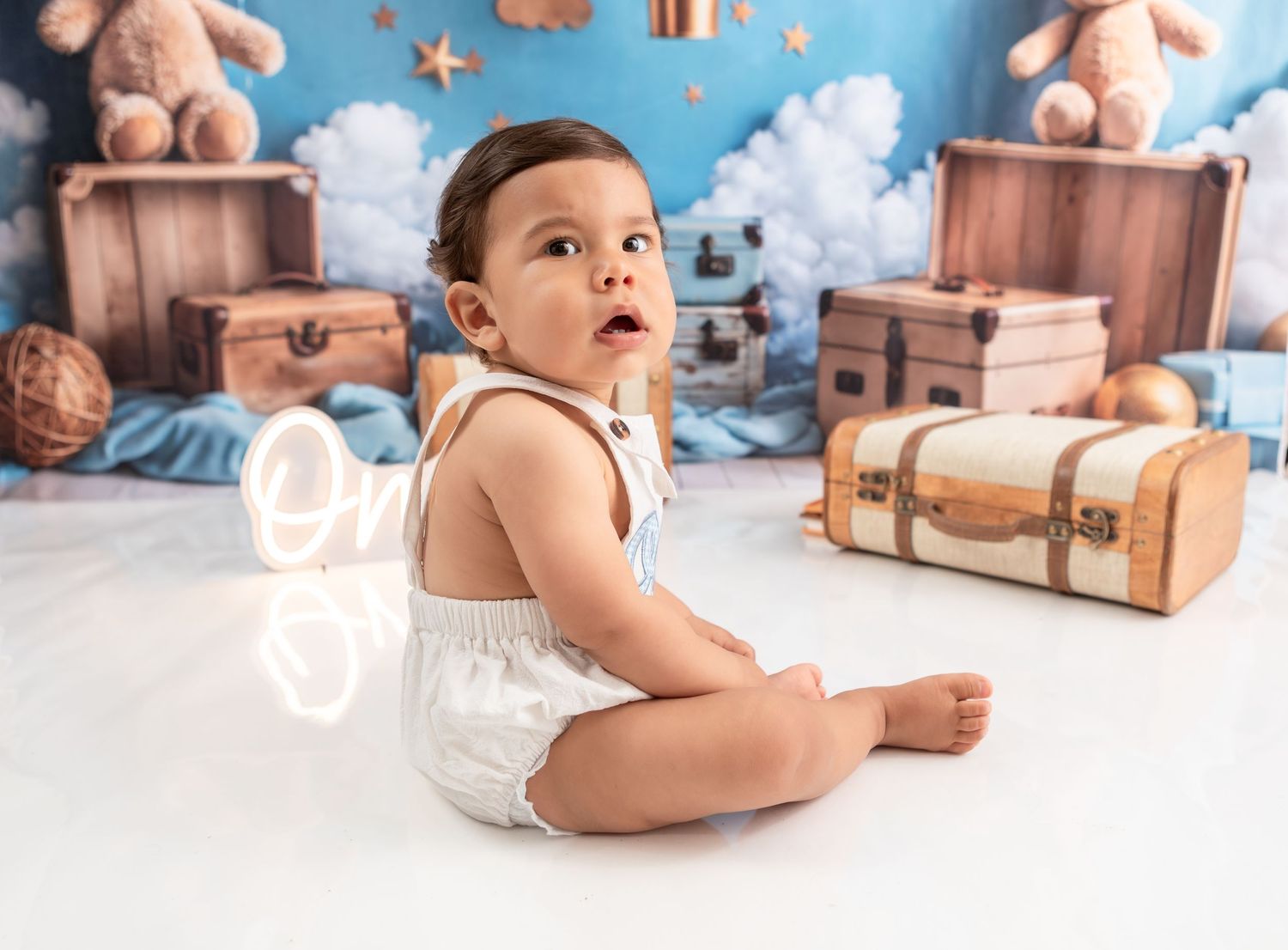 Baby in white onesie sitting against travel-themed backdrop with suitcases and clouds.